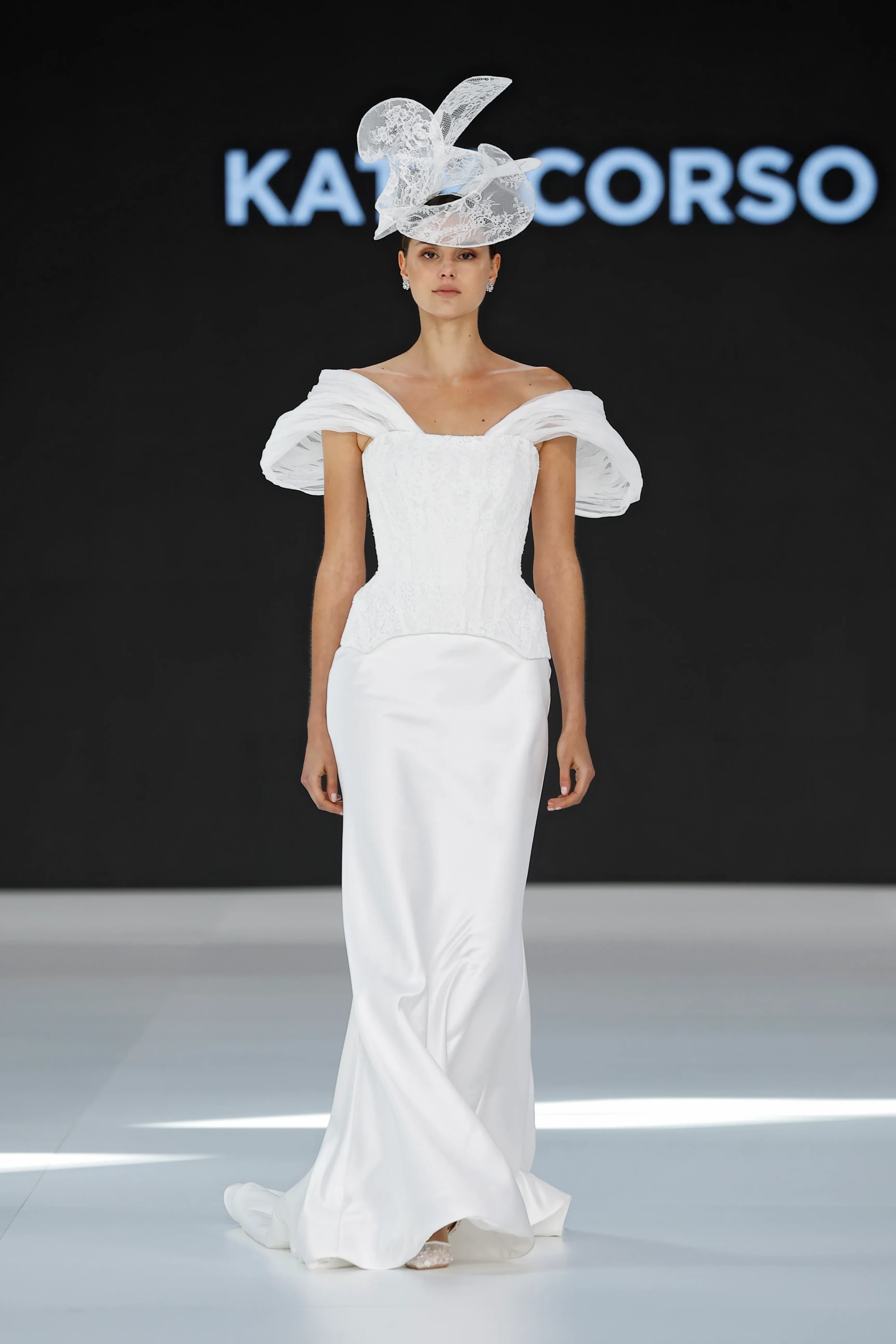 Barcelona Bridal Fashion Week 2026 podyumunda, Katy Corso İlkbahar 2027 Gelinlik Koleksiyonu'ndan omuzları açık, korse detaylı ve kabarık tül kollu, akışkan saten etekli beyaz bir gelinlik giyen model, başındaki heykelsi dantel aksesuarla hafiflik ve couture hassasiyetini zarifçe sergiliyor.