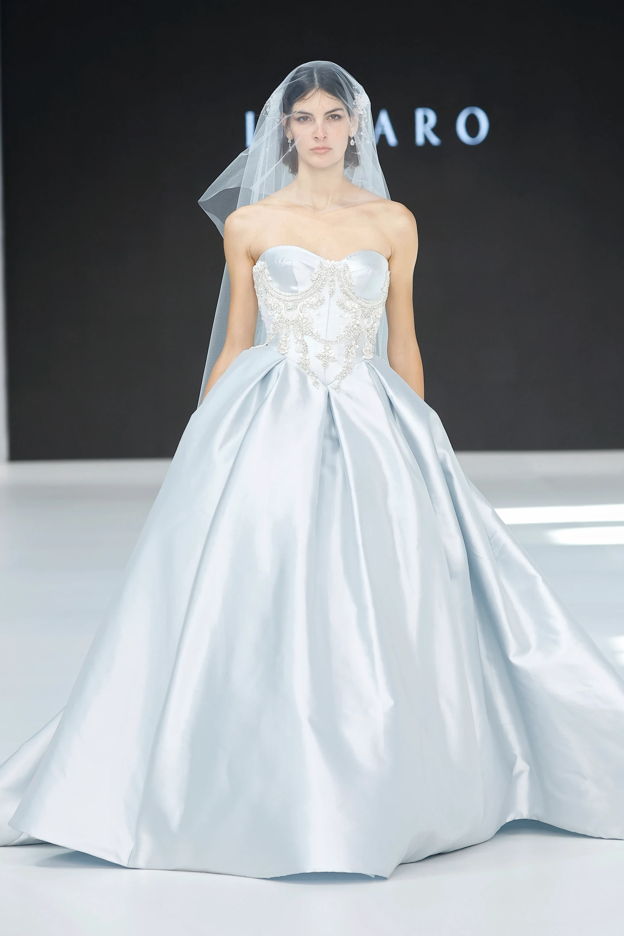 Barcelona Bridal Fashion Week 2026'da sergilenen, straplez gümüşi mavi, üst kısmı zarif işlemelerle bezeli korsaj detayına sahip, hacimli etekli Lazaro gelinlik modeli sade duvağıyla 2027 gelinlik trendlerini yansıtarak podyumda romantik bir siluet sunuyor.