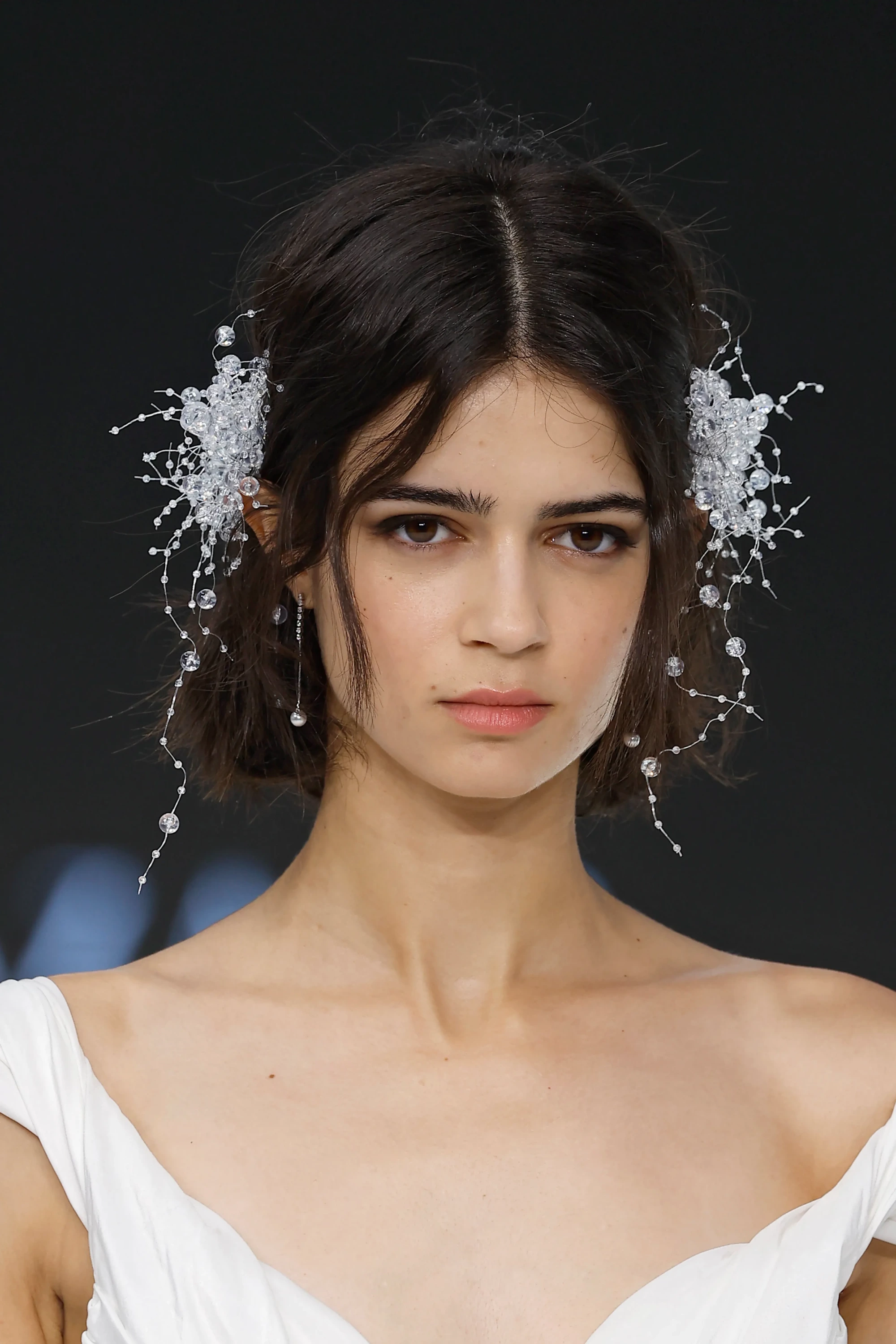 Barcelona Bridal Fashion Week 2026’da Lazaro & Maison Pérez 2027 İlkbahar Gelinlik Koleksiyonu’ndan, omuzları açık beyaz bir gelinlik giyen modelin, modern ve romantik bir gelinlik trendini temsil eden ışıltılı kristal saç aksesuarlarıyla süslenmiş yakın çekim portresi.