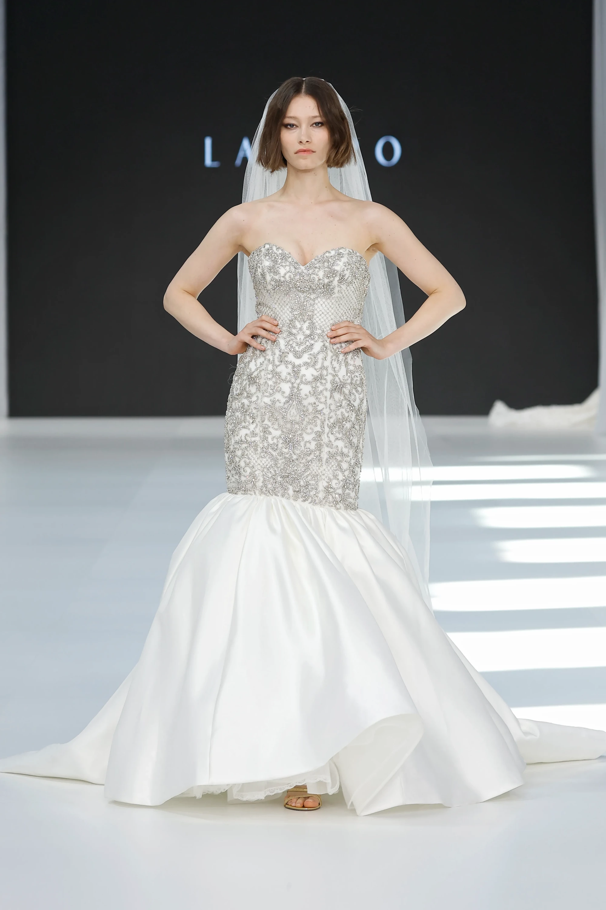 Barcelona Bridal Fashion Week 2026 podyumunda kısa saçlı bir model, 2027 gelinlik trendlerini yansıtan, üst kısmı gümüş işlemelerle bezeli straplez, denizkızı kesim ve hacimli saten etekli, modern lüksü romantik bir zarafetle birleştiren bir Lazaro gelinliği ve zarif bir duvakla poz veriyor.