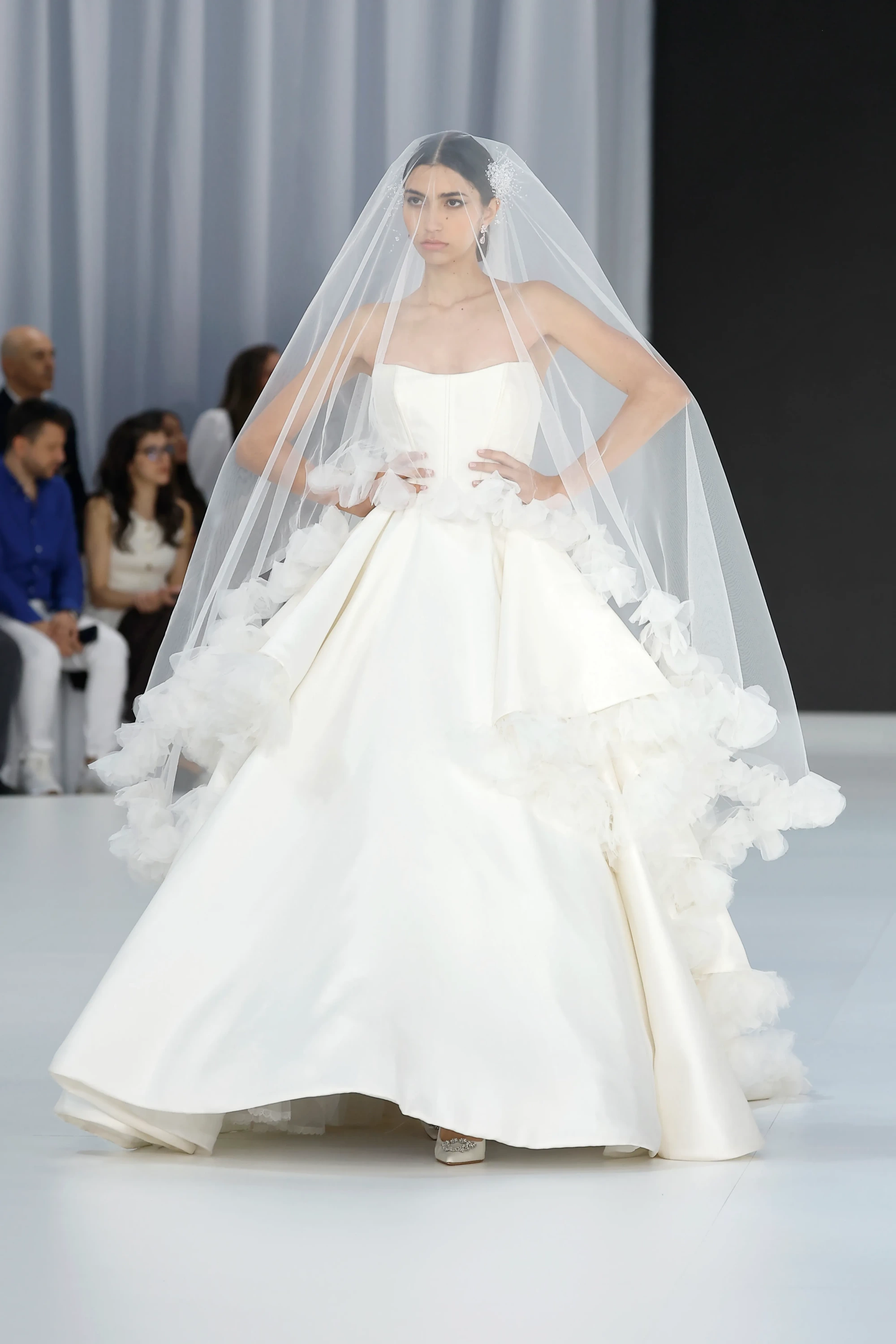 Pistte elleri belinde duran bir model, modern lüksü romantik zarafetle harmanlayan, straplez, hacimli saten eteği ve bel kısmındaki kat kat fırfırlı tül detaylarıyla dikkat çeken bembeyaz bir gelinliği uzun, şeffaf duvağıyla birlikte sergileyerek 2027 gelinlik trendlerine göz kırpıyor.