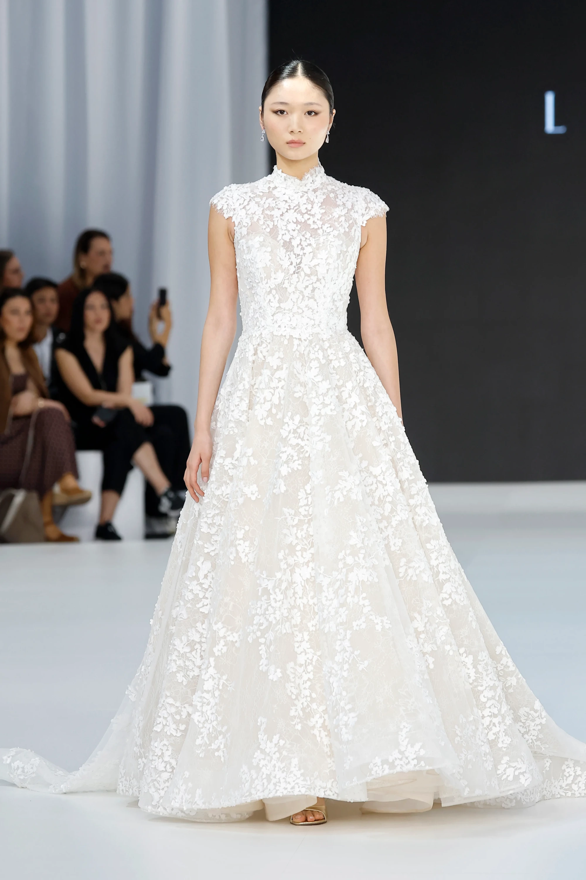 Barcelona Bridal Fashion Week 2026'da podyumda yürüyen model, Lazaro & Maison Pérez imzalı, yüksek yakalı, kabartmalı çiçek desenleriyle süslenmiş, hacimli etekli, zarif bir gelinlik giyiyor.