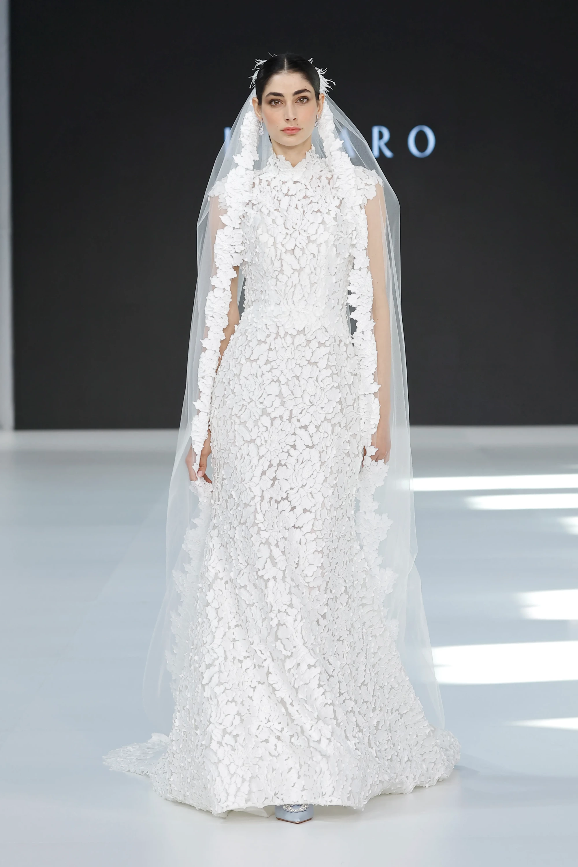 Barcelona Bridal Fashion Week 2026'dan: Yüksek yakalı, bedeni saran, tamamı yoğun 3D çiçek aplikeli beyaz gelinliği ve kenarları benzer çiçeklerle süslü uzun, modern duvağıyla podyumda yürüyen model, 2027 gelinlik trendlerinden modern duvak ve 3D çiçeklerin güçlü yorumunu gözler önüne seriyor.