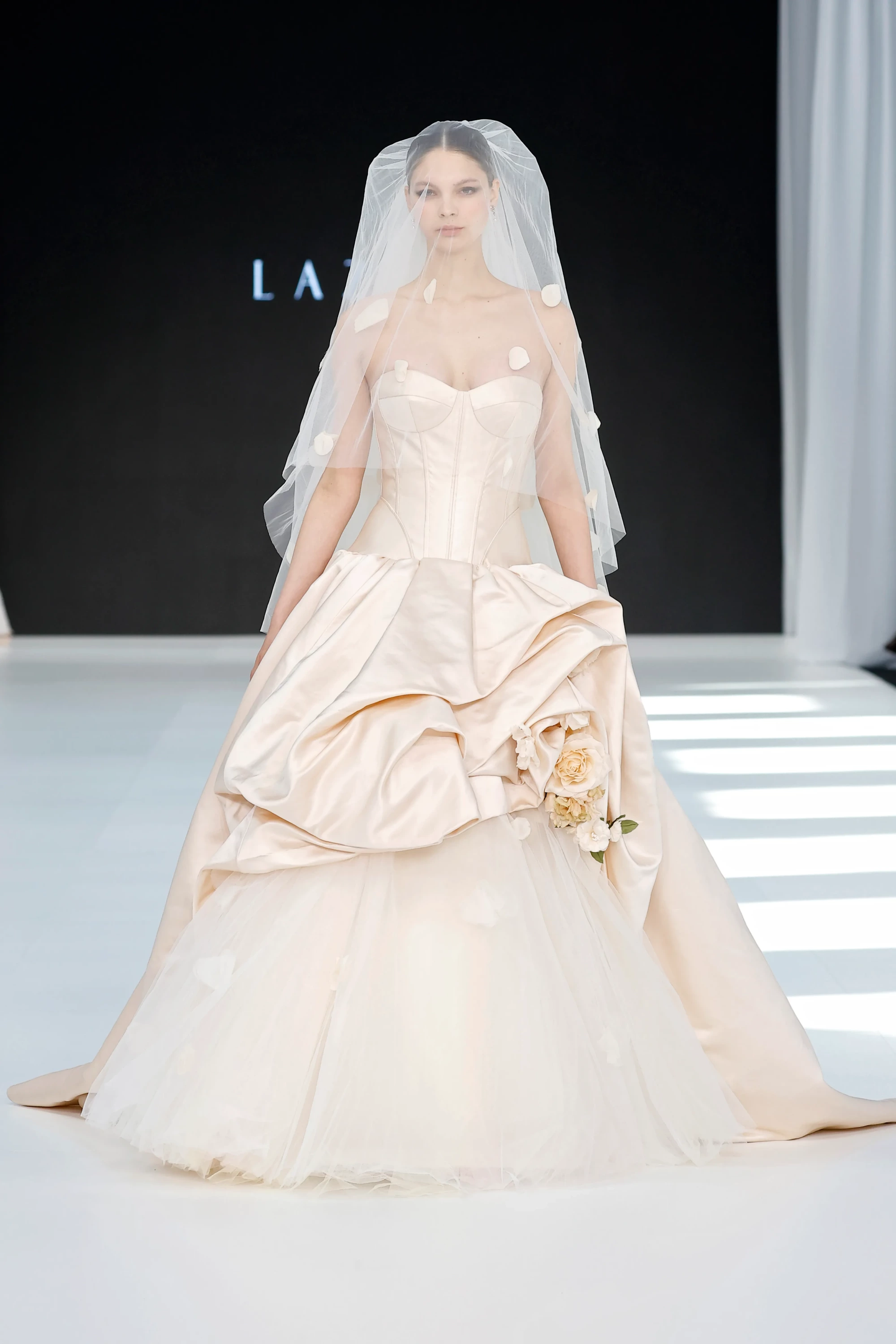 Lazaro & Maison Pérez'in 2027 İlkbahar Gelinlik Koleksiyonu'ndan, podyumda düz duran model, balerin etkisi taşıyan, kat kat tül etekli, korse üst detaylı ve belinde romantik çiçek aplikeleri bulunan şampanya rengi göz alıcı bir gelinlik giyiyor. Bu gelinlik, 2027 gelinlik trendleri arasında yer alan hacimli ve hikâyesi olan siluetlere vurgu yapıyor.