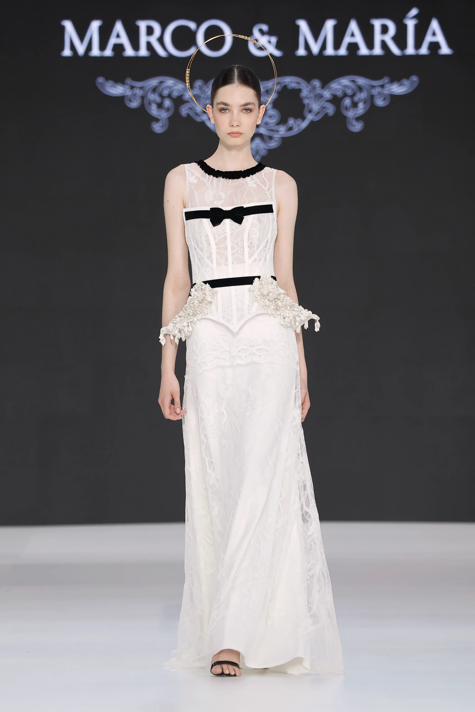 Barcelona Bridal Fashion Week 2026 kapsamında sergilenen Marco & María 2027 İlkbahar Gelinlik Koleksiyonu'ndan, zarif altın rengi hale başlığı, siyah kadife yaka ve fiyonk detayları, beldeki üç boyutlu çiçek aplikasyonları ve akışkan dantel eteğiyle modern bir gelinlik giyen model, 2027 gelinlik modasının çağdaş yorumunu sergiliyor.