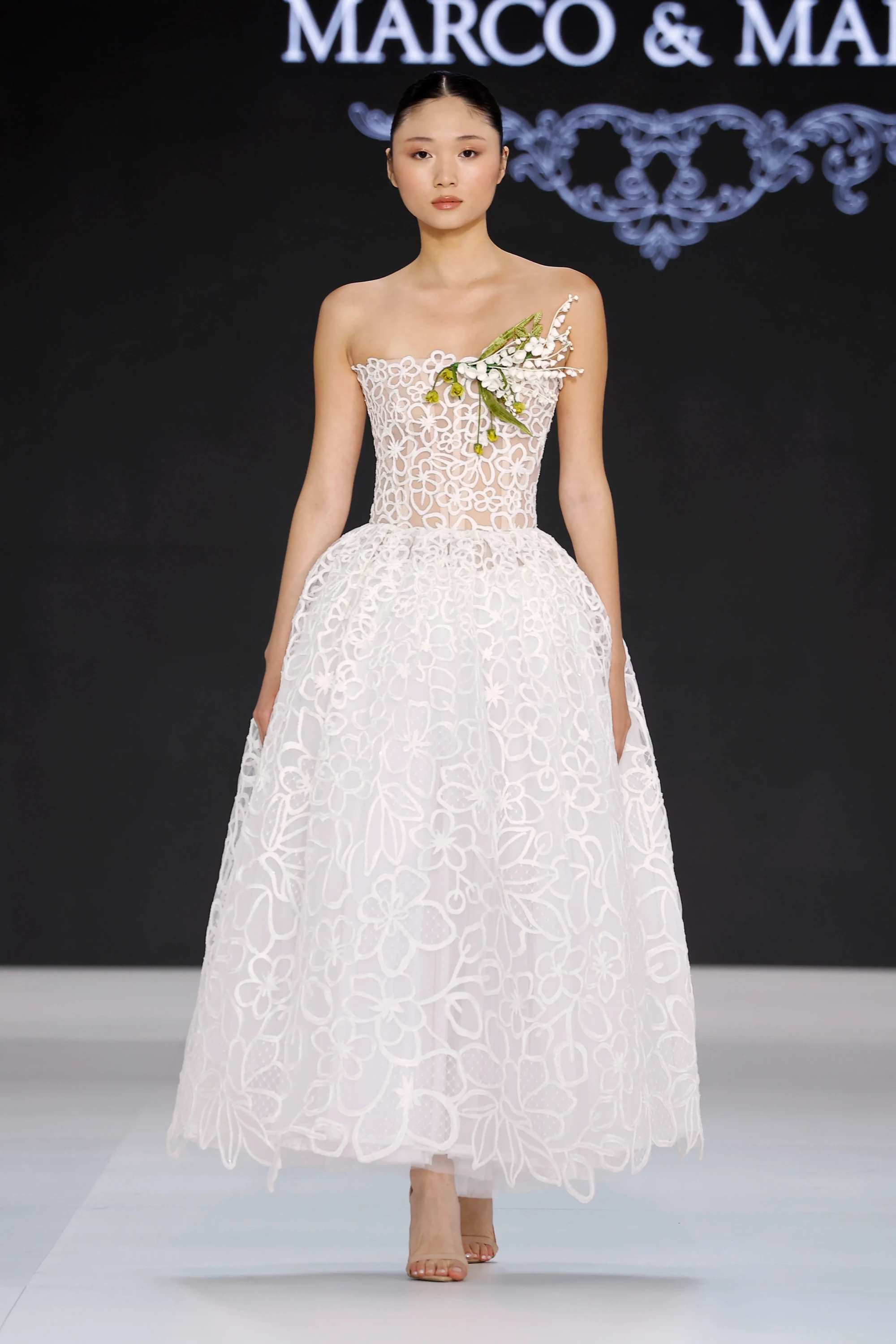 Barcelona Bridal Fashion Week'te sergilenen Marco & María 2027 İlkbahar Gelinlik Koleksiyonu'ndan, göğsünde doğal yeşil yapraklar ve beyaz çiçeklerden oluşan 3D aplikelerle süslenmiş, transparan korsajlı, zarif dantel işlemeli ve hafif hacimli, midi boy beyaz bir gelinlik model üzerinde sunuluyor.