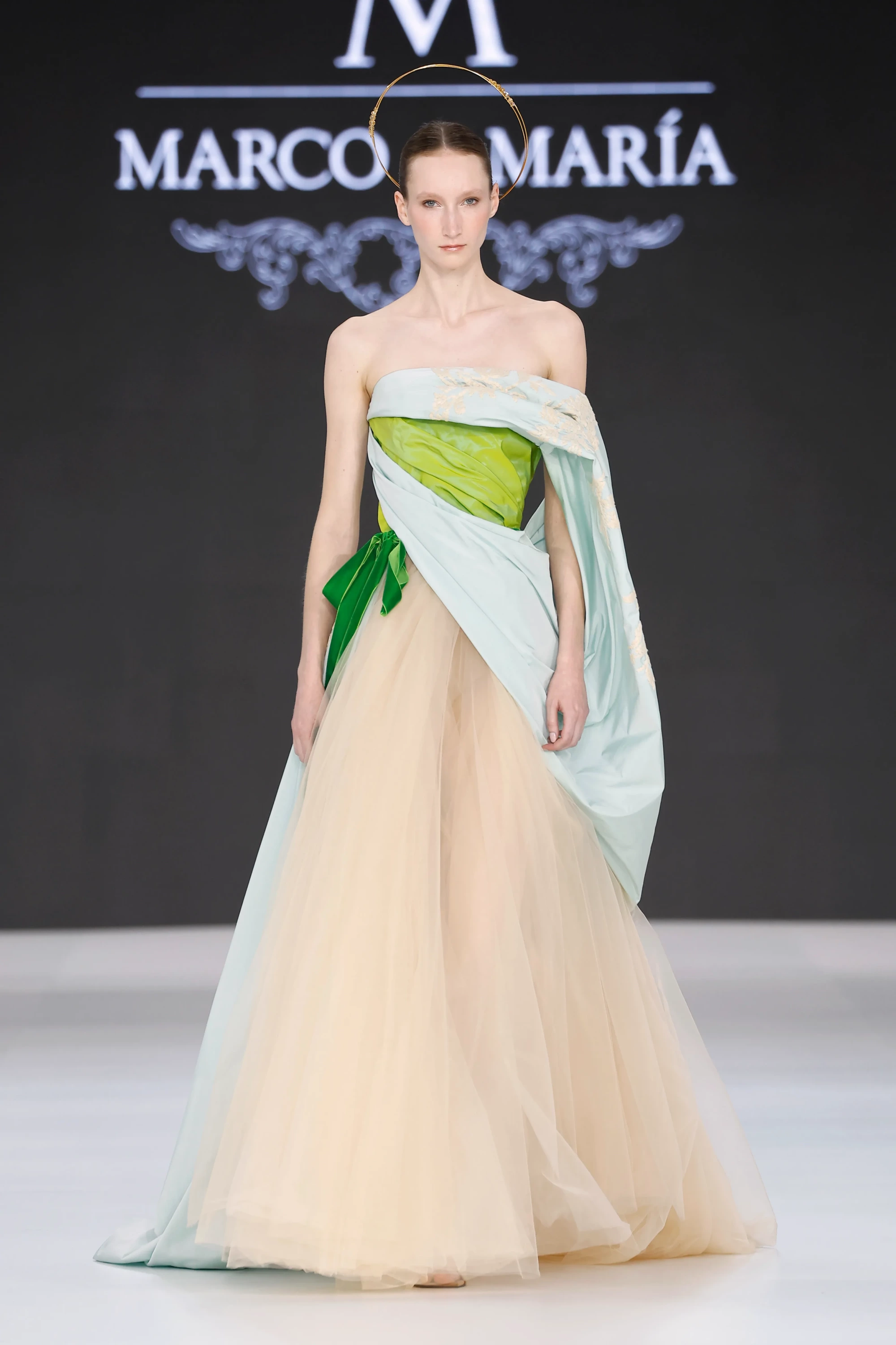 Bir model, üstü canlı yeşil ve mint yeşili nakışlı drape kumaşla sarılı, bej tül etekli straplez bir gelinlik giymiş, saçında altın rengi haleli bir aksesuarla podyumda duruyor; bu tasarım Marco & María'nın Barcelona Bridal Fashion Week'te sergilediği 2027 İlkbahar gelinlik koleksiyonundan modern romantizmi yansıtan katmanlı ve zarif bir örnek.