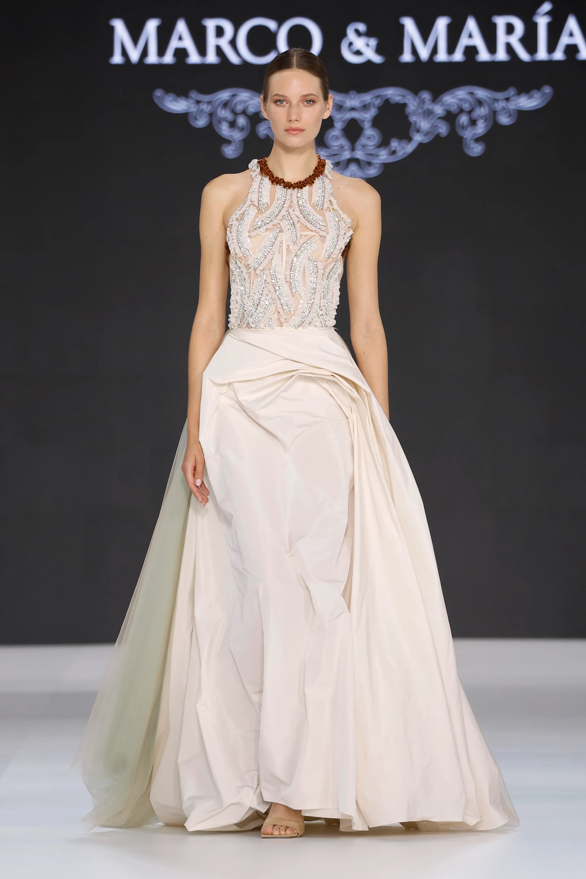 Marco & María'nın Barcelona Bridal Fashion Week'te sergilenen 2027 İlkbahar Gelinlik Koleksiyonu'ndan, boyun kısmında kahverengi fırfırlı detayla zenginleşen, gümüş boncuk işlemeli transparan halter yakalı üst kısmı ve beli vurgulayan hacimli, dökümlü eteğiyle modern romantizmi yansıtan katmanlı bir gelinlik giyen model podyumda.