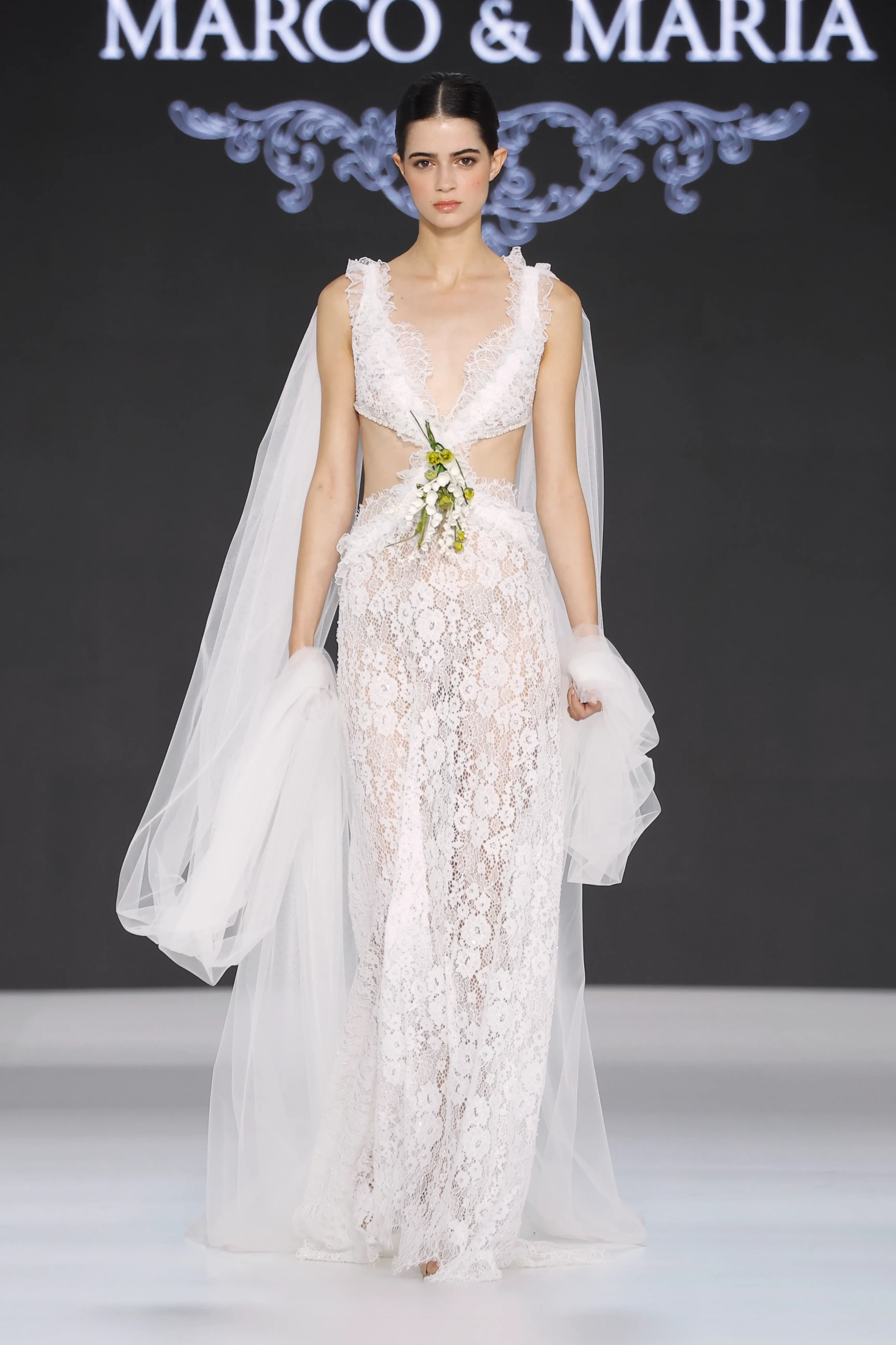 Barcelona Bridal Fashion Week'te Marco & María'nın 2027 İlkbahar Gelinlik Koleksiyonu'ndan, yanları transparan kesik dekolteli ve ortasında yeşil-beyaz çiçek detayları bulunan, tümü zarif dantel işlemeli, omuzlarından yere kadar uzanan uçuşan tül pelerinli bir gelinliği tanıtan model podyumda zarifçe yürüyor.