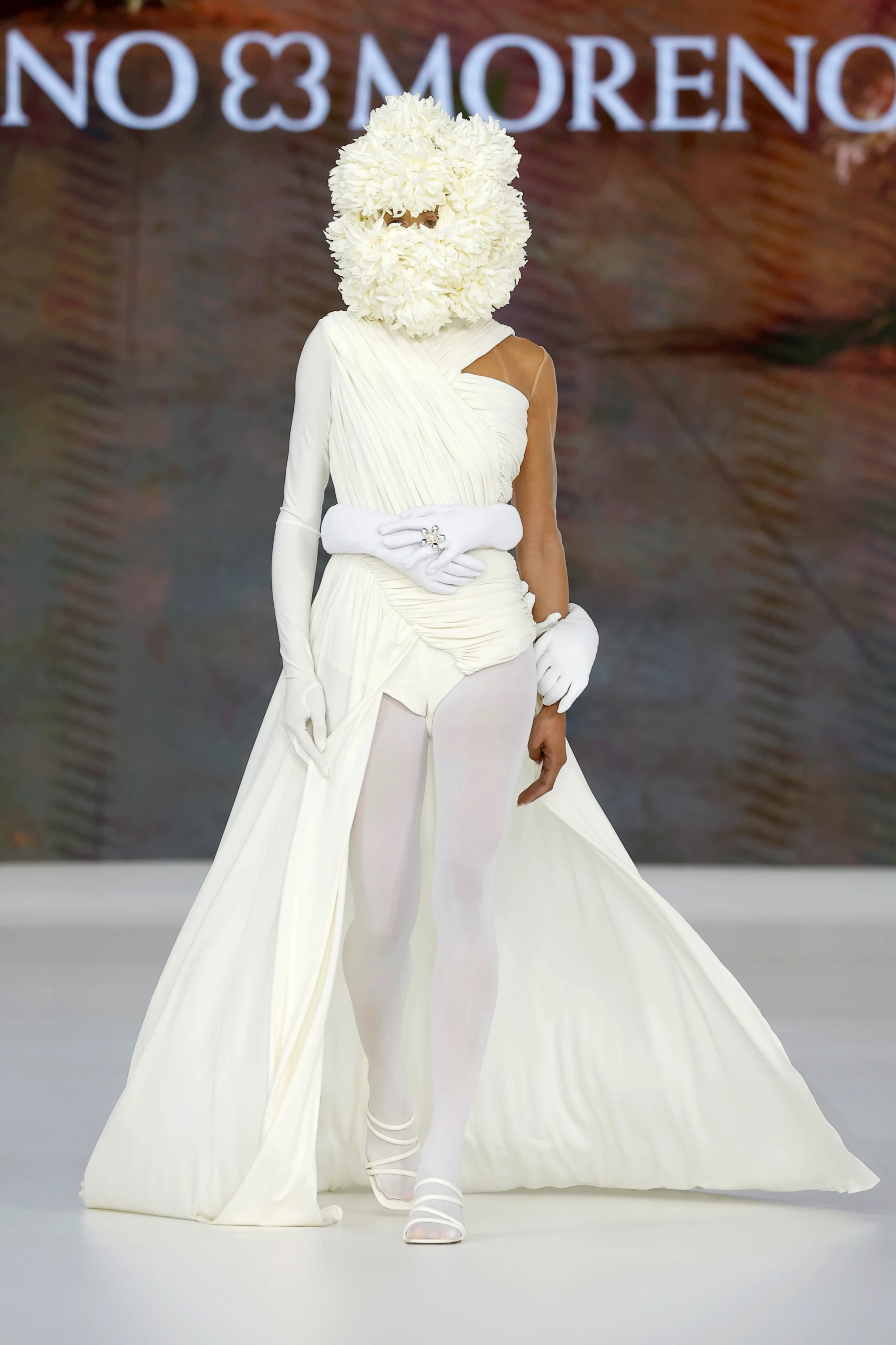 Mariano Moreno'nun Barcelona Bridal Fashion Week 2026'da sergilenen 2027 İlkbahar gelinlik koleksiyonundan bir model, yüzünü gizleyen büyük beyaz çiçek maskesi, tek omuzlu ve derin yırtmaçlı drapeli beyaz elbisesi, belindeki eldivenli eller ve pırıltılı yüzük detayıyla botanik dünyadan ilham alan giyilebilir bir sanat eseri sergiliyor.
