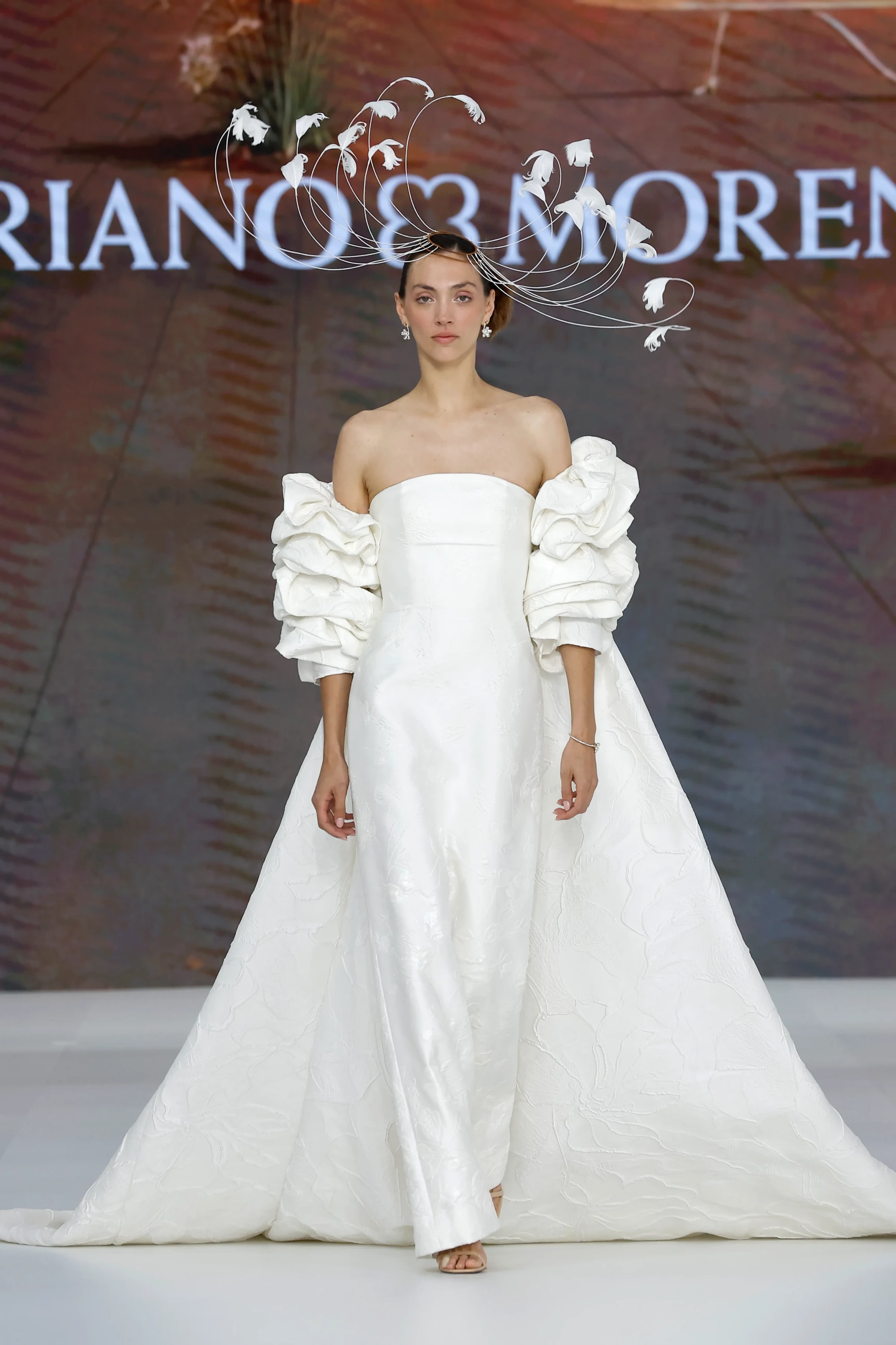 Barcelona Bridal Fashion Week podyumunda, Mariano Moreno'nun 2027 İlkbahar gelinlik koleksiyonundan, hacimli kolları ve bitkisel detaylarla süslenmiş heykelsi bir başlığı olan straplez beyaz gelinliği tanıtan bir model görünüyor, bu tasarım koleksiyonun sanatsal ve yenilikçi duruşunu yansıtıyor.