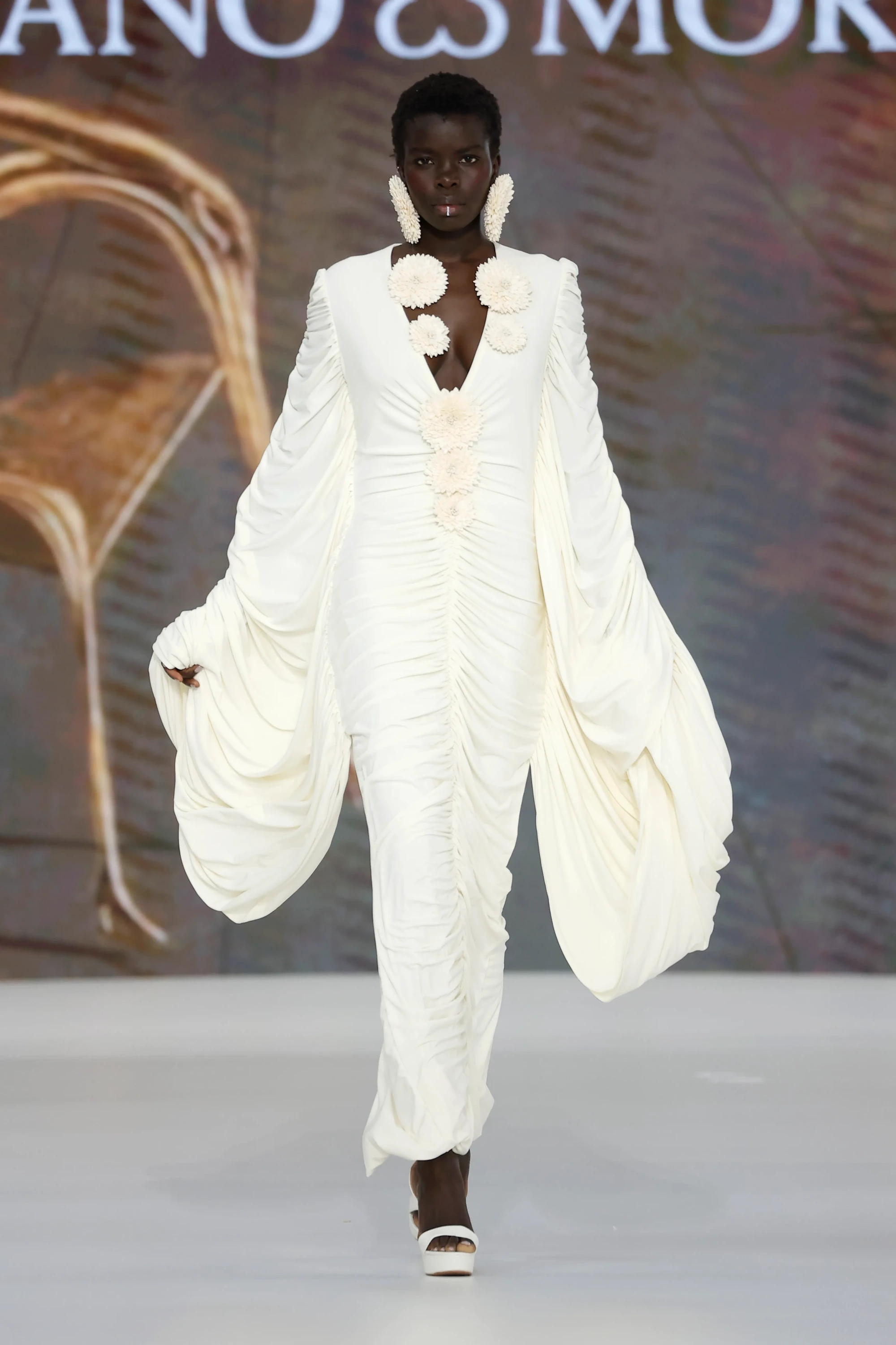 Barcelona Bridal Fashion Week'te Mariano Moreno'nun 2027 İlkbahar gelinlik koleksiyonundan, botanik dünyadan ilham alan, derin V yakalı, hacimli balon kollu ve göğüs ile eteğinde yapısal üç boyutlu çiçek aplikeleriyle süslenmiş, tüm vücudu saran büzgülü ekru bir gelinlik giyen model, Rafa Peinador imzalı büyük çiçek küpeleriyle podyumda göz alıcı bir duruş sergiliyor.