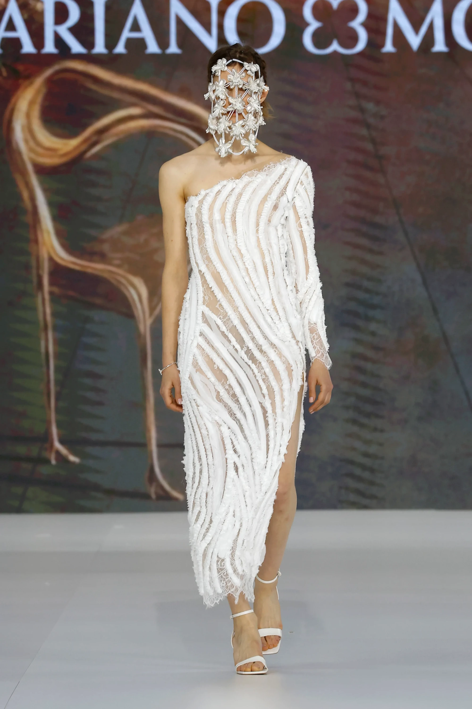 Mariano Moreno'nun Barcelona Bridal Fashion Week 2026 defilesinde, botanik ilhamlı, yüzü saran üç boyutlu çiçek formunda başlığı ve asimetrik kesimli, dokulu beyaz gelinliği ile podyumda yürüyen bir model, 2027 gelinlik trendlerine iddialı bir bakış sunuyor.