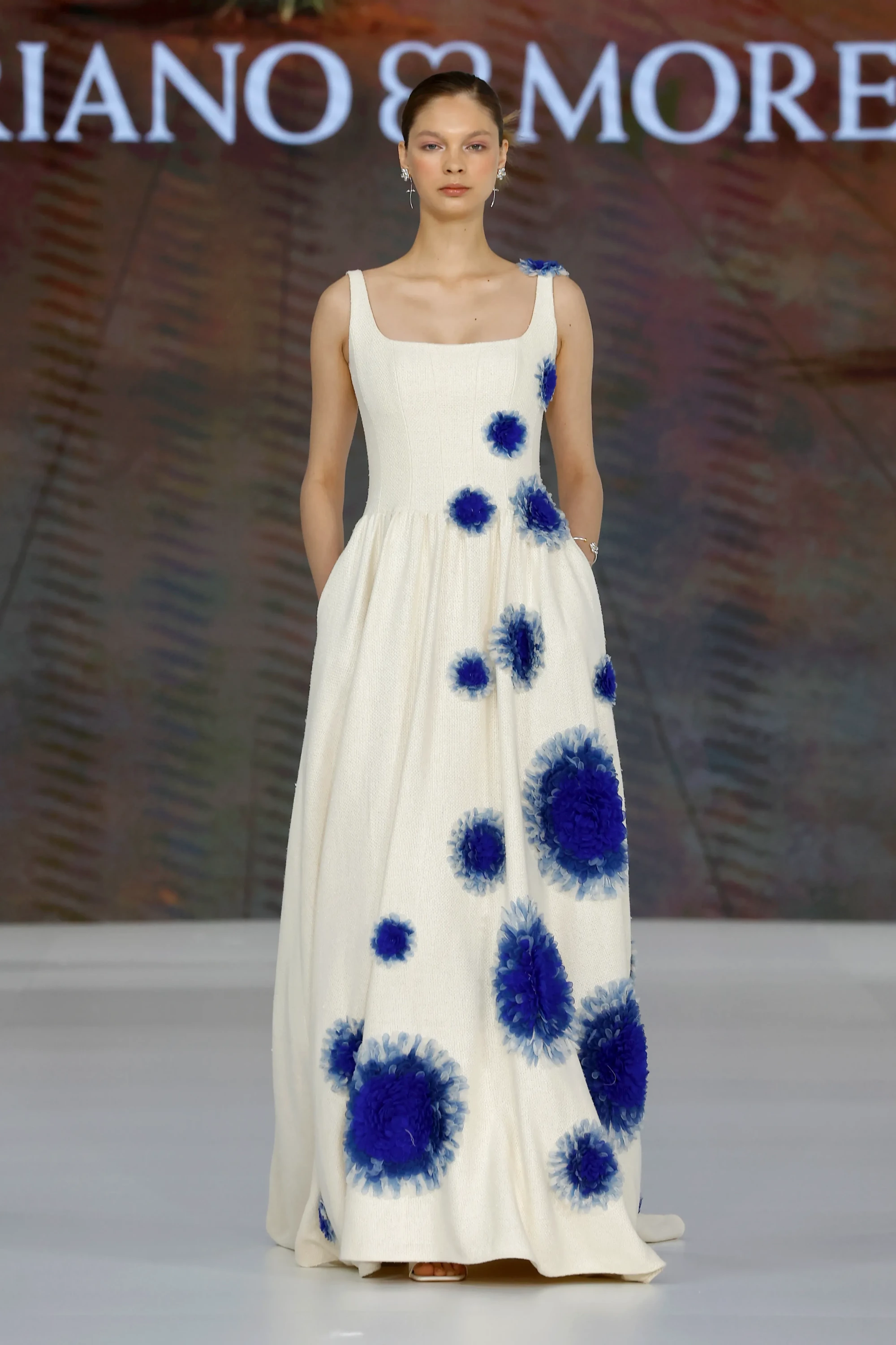 Mariano Moreno'nun Barcelona Bridal Fashion Week 2026 defilesinden, 2027 gelinlik trendleri arasında öne çıkan, vücudu saran kare yaka üst kısmı ve cepli dökümlü eteğiyle dikkat çeken ekru rengi gelinlik, yüzeyine serpiştirilmiş canlı mavi tonlardaki hacimli 3D çiçek aplikeleriyle botanik bir zarafet sunuyor.