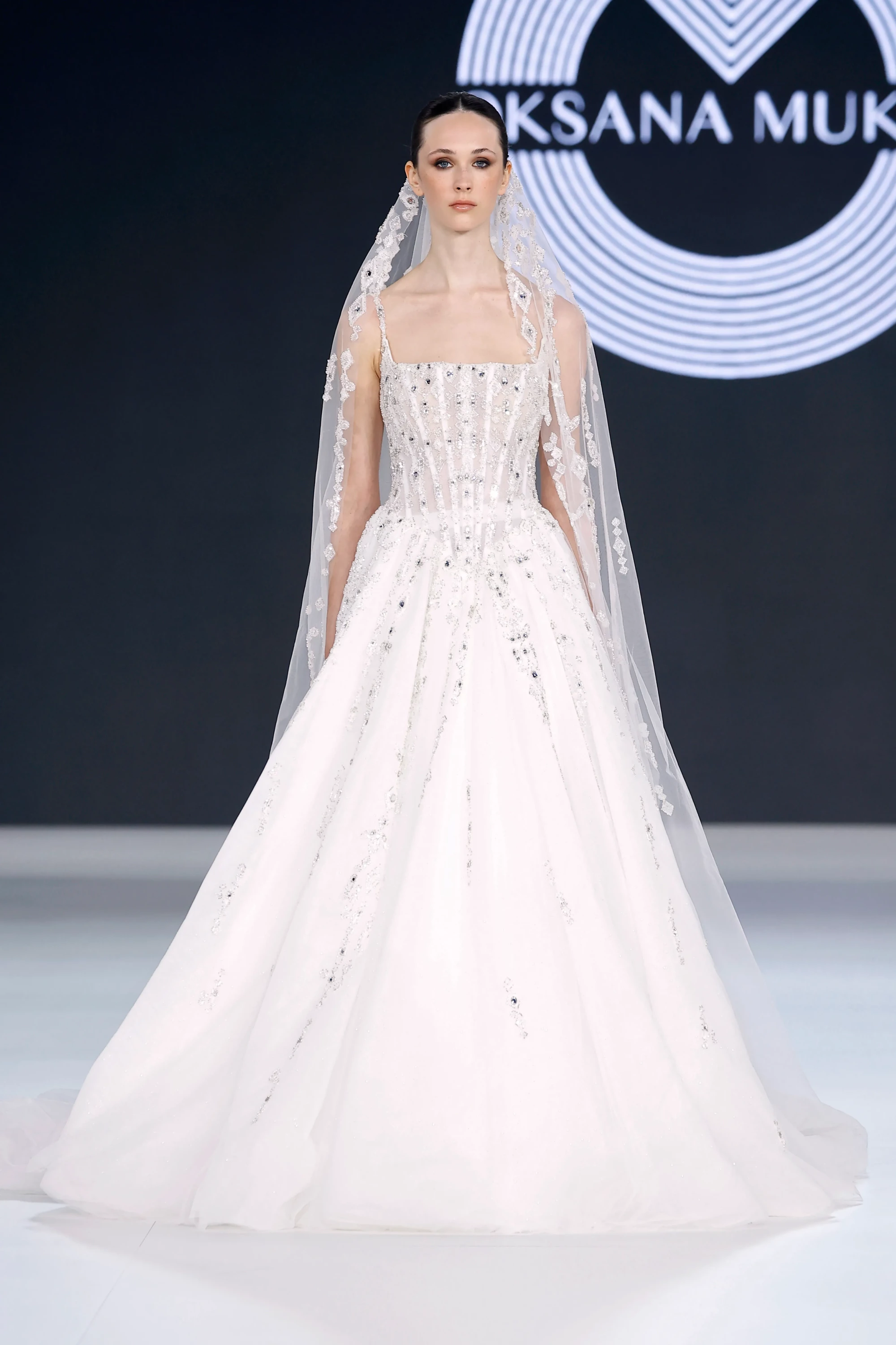Oksana Mukha'nın Barcelona Bridal Fashion Week 2026'da sunduğu 2027 İlkbahar gelinlik koleksiyonundan, işlemeli kare yaka korsajlı üst kısmı, kat kat tül eteği ve ışıltılı uzun duvağıyla podyumda duran model, modern romantizm ve haute couture ustalığını yansıtıyor.