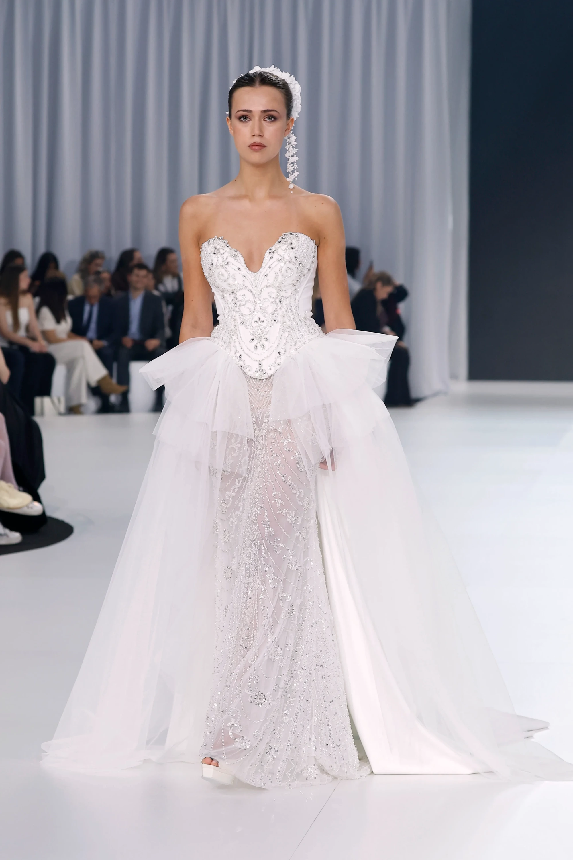 Barcelona Bridal Fashion Week 2026'da Oksana Mukha'nın 2027 İlkbahar gelinlik koleksiyonundan, modern romantizm ve el işçiliği işlemelerle öne çıkan, straplez, korse etkili büstiyeri ve belinde hafif hacimli tül peplum detayı bulunan, pırıl pırıl boncuk işlemeli transparan eteği ve akışkan uzun kuyruğuyla dikkat çeken bir gelinlik podyumda sergileniyor.