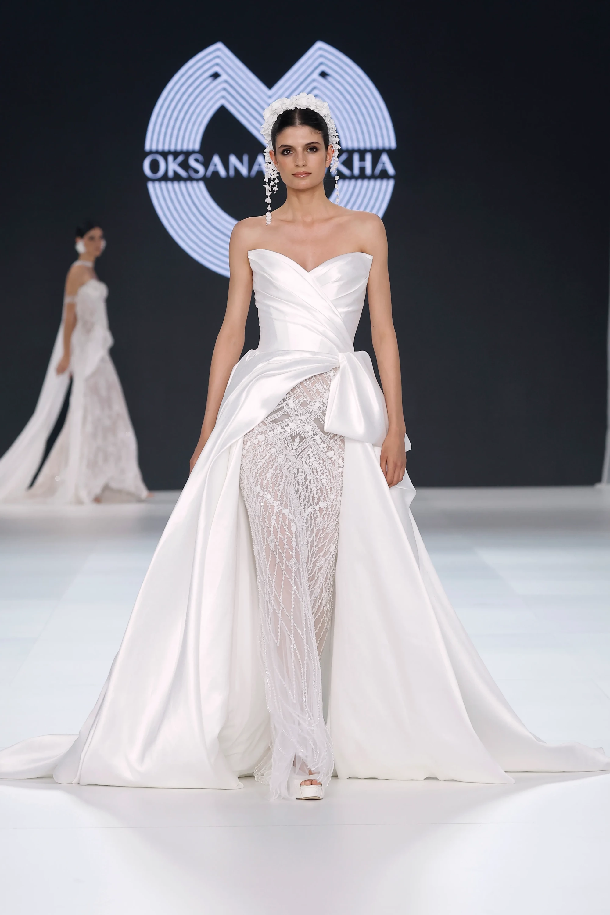 Oksana Mukha'nın 2027 İlkbahar gelinlik koleksiyonundan, podyumda yürüyen modelin üzerindeki straplez, korsajlı üst kısmı ve önü açılan kabarık saten eteği, altındaki ışıltılı işlemeli dar silüetle modern romantizmi harmanlayan tasarımı yansıtırken, başında ve sallantılı küpelerinde floral aplikeler göze çarpıyor.
