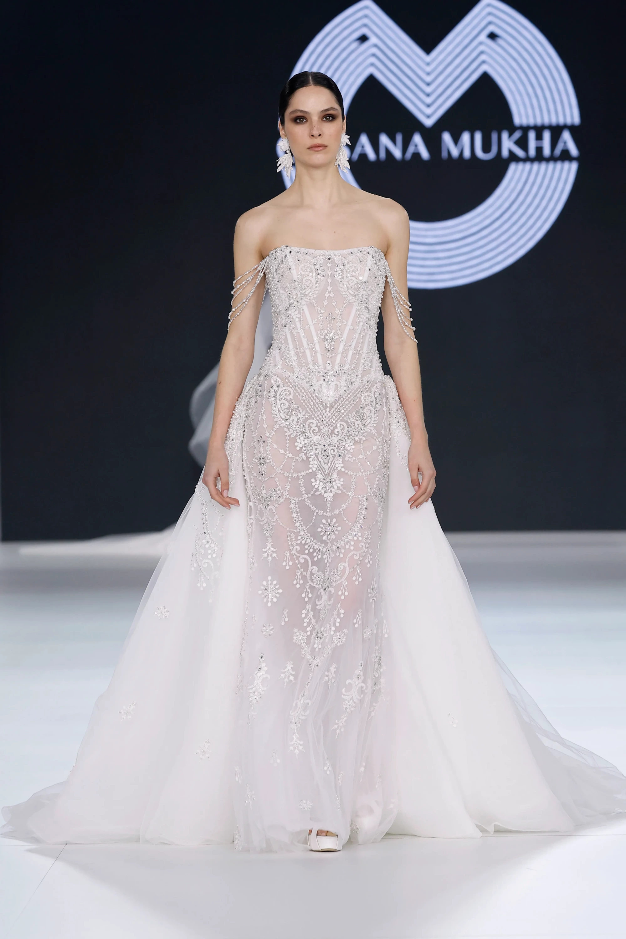 Podyumda yürüyen bir model, Barcelona Bridal Fashion Week'te sergilenen Oksana Mukha'nın 2027 İlkbahar Gelinlik Koleksiyonu'ndan, vücudu saran, straplez, incilerle işlenmiş, şeffaf, uzun kuyruklu ve omuzlardan sarkan boncuk zincir detaylı, modern romantizmi yansıtan bir gelinlik giyiyor.