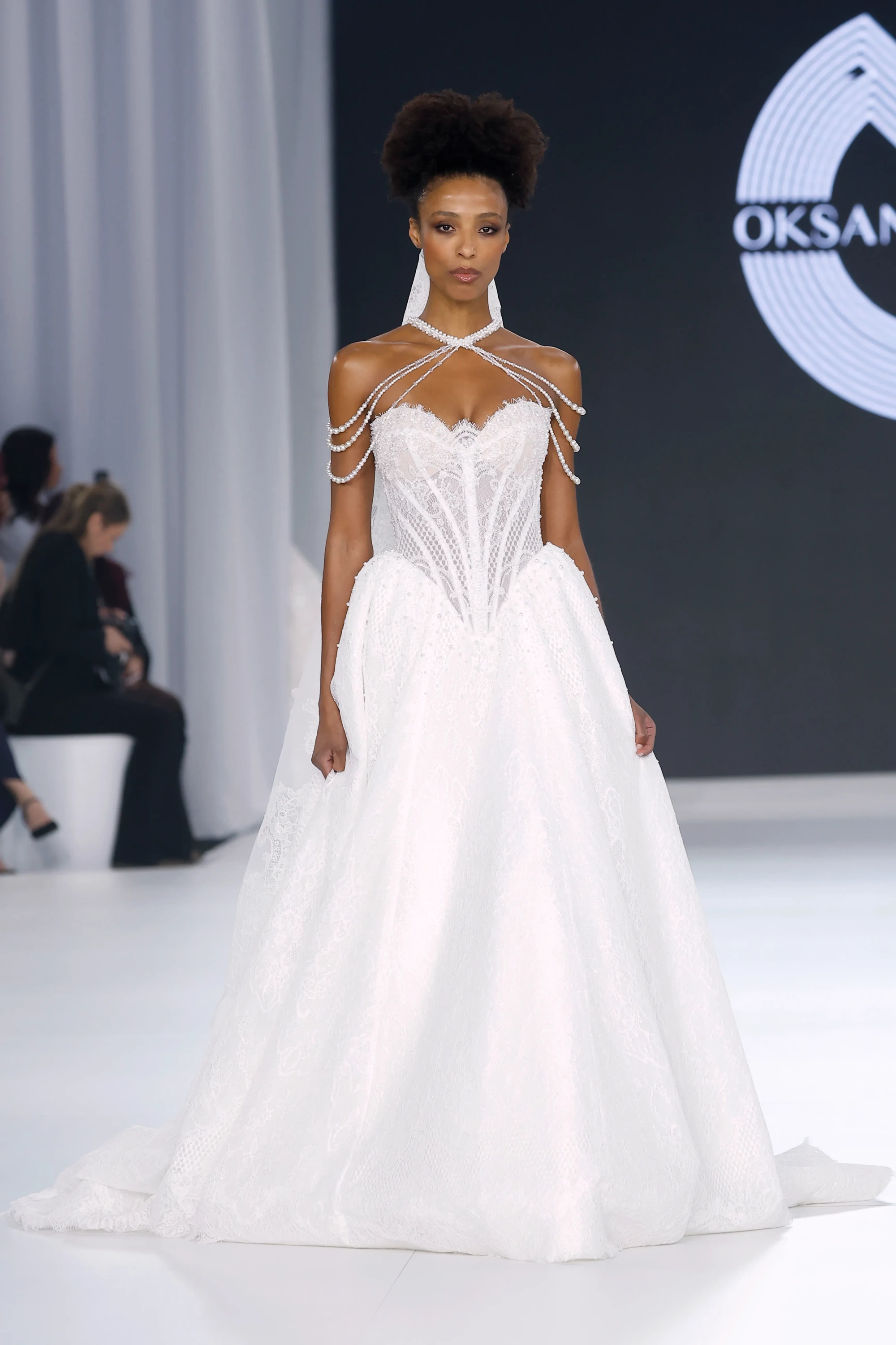 Oksana Mukha'nın Barcelona Bridal Fashion Week'te sergilenen 2027 İlkbahar gelinlik koleksiyonundan, incilerle zenginleştirilmiş omuz askıları, dantel işlemeli korse büstiyeri ve katmanlı kabarık eteğiyle dikkat çeken beyaz bir gelinlik giyen model, podyumda zarifçe süzülüyor.