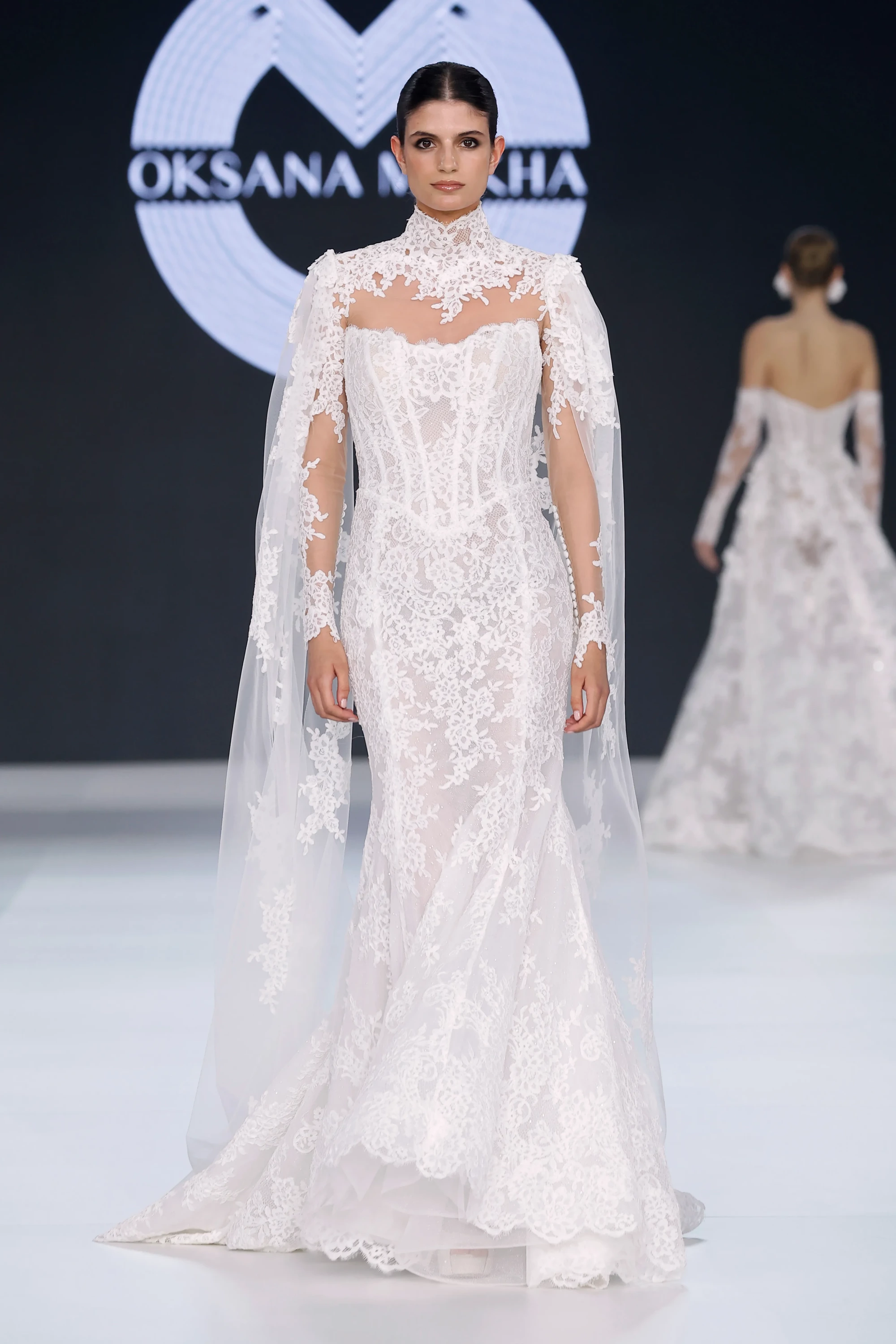 Oksana Mukha'nın Barcelona Bridal Fashion Week 2026'da sunduğu 2027 ilkbahar gelinlik koleksiyonundan, yüksek yakalı, detaylı dantel işlemeli, transparan uzun kollu ve omuzlardan zarifçe sarkan pelerin detaylarına sahip, modern romantizm ve Viktoryen dokunuşları harmanlayan balık etekli bir gelinlik giymiş model podyumda yürüyor.