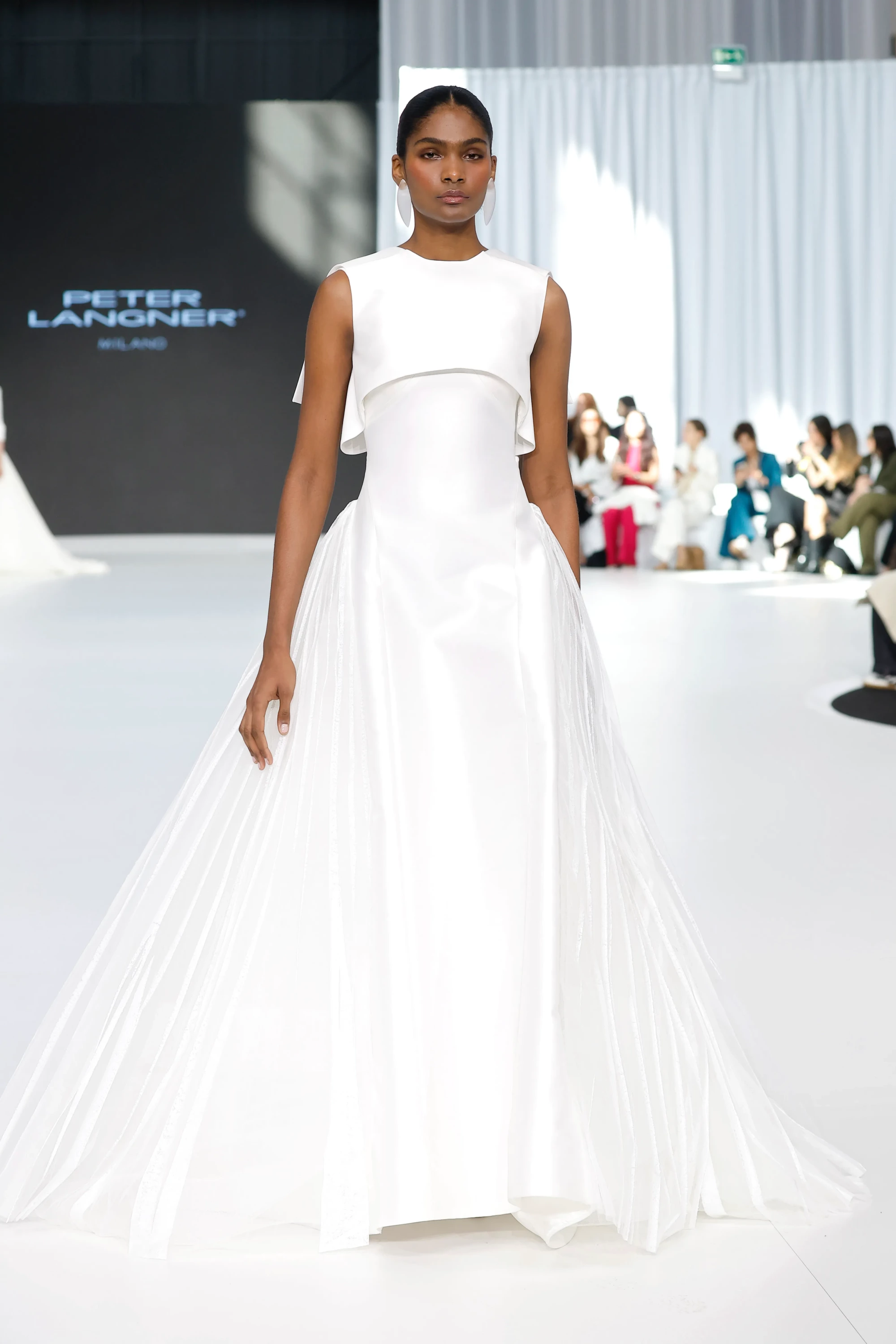 Peter Langner'ın Barcelona Bridal Fashion Week 2026 defilesinden, omuzları kısa pelerin detaylı, bedeni saran saten ve alt kısmı katmanlı, hacimli tülden oluşan, 2027 gelinlik trendlerini yansıtan modern beyaz bir gelinlik modeli, podyumda zarif bir şekilde sunuluyor.