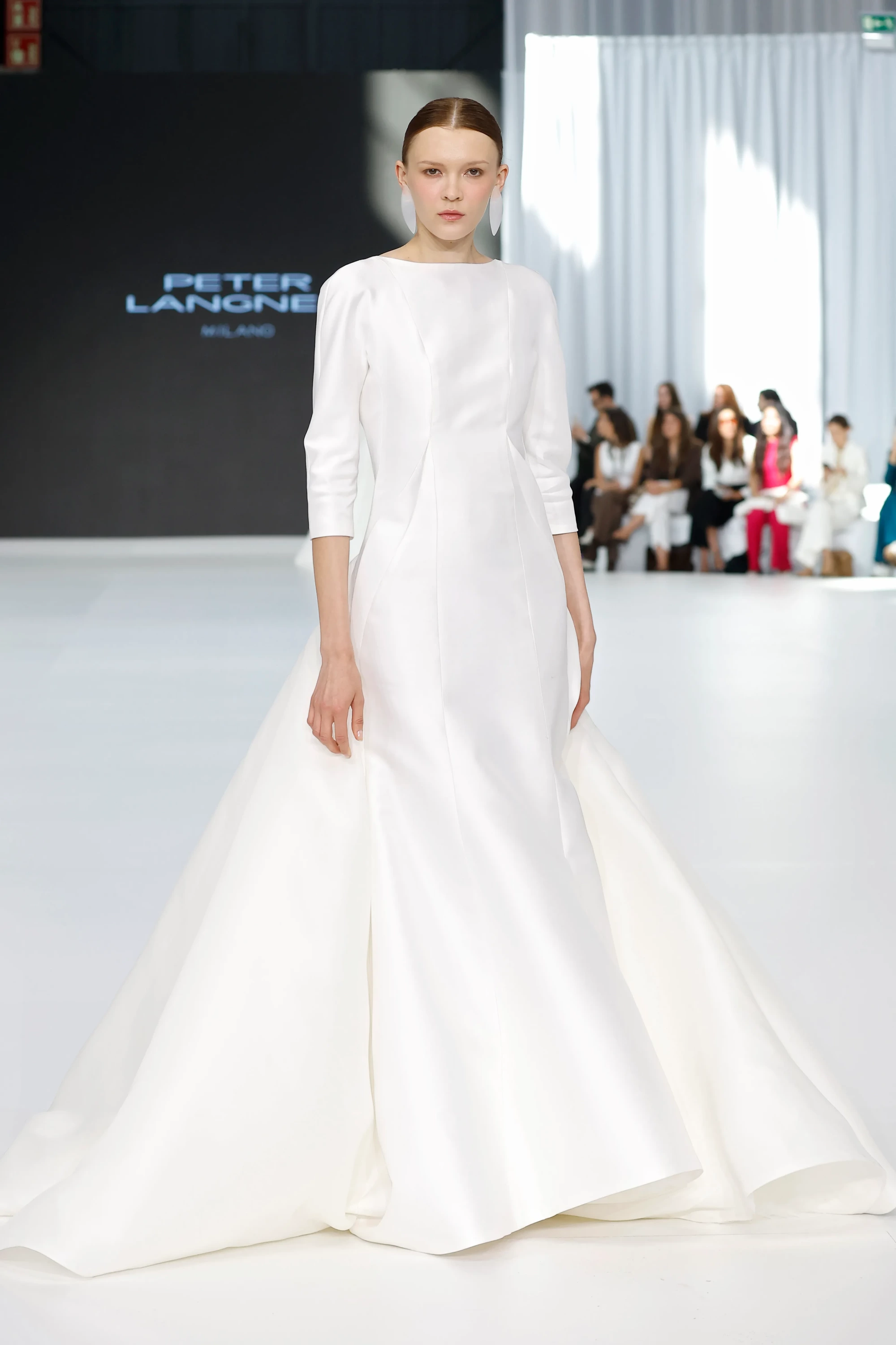 Peter Langner'in 2026 Barcelona Bridal Fashion Week defilesinden, yüksek yuvarlak yakalı, dirsekte biten puf kollu ve hacimli A kesim etekli, mat beyaz mikado kumaştan zarif bir gelinlik giyen model, minimalist beyaz küpelerle 2027 gelinlik trendlerini sunuyor.