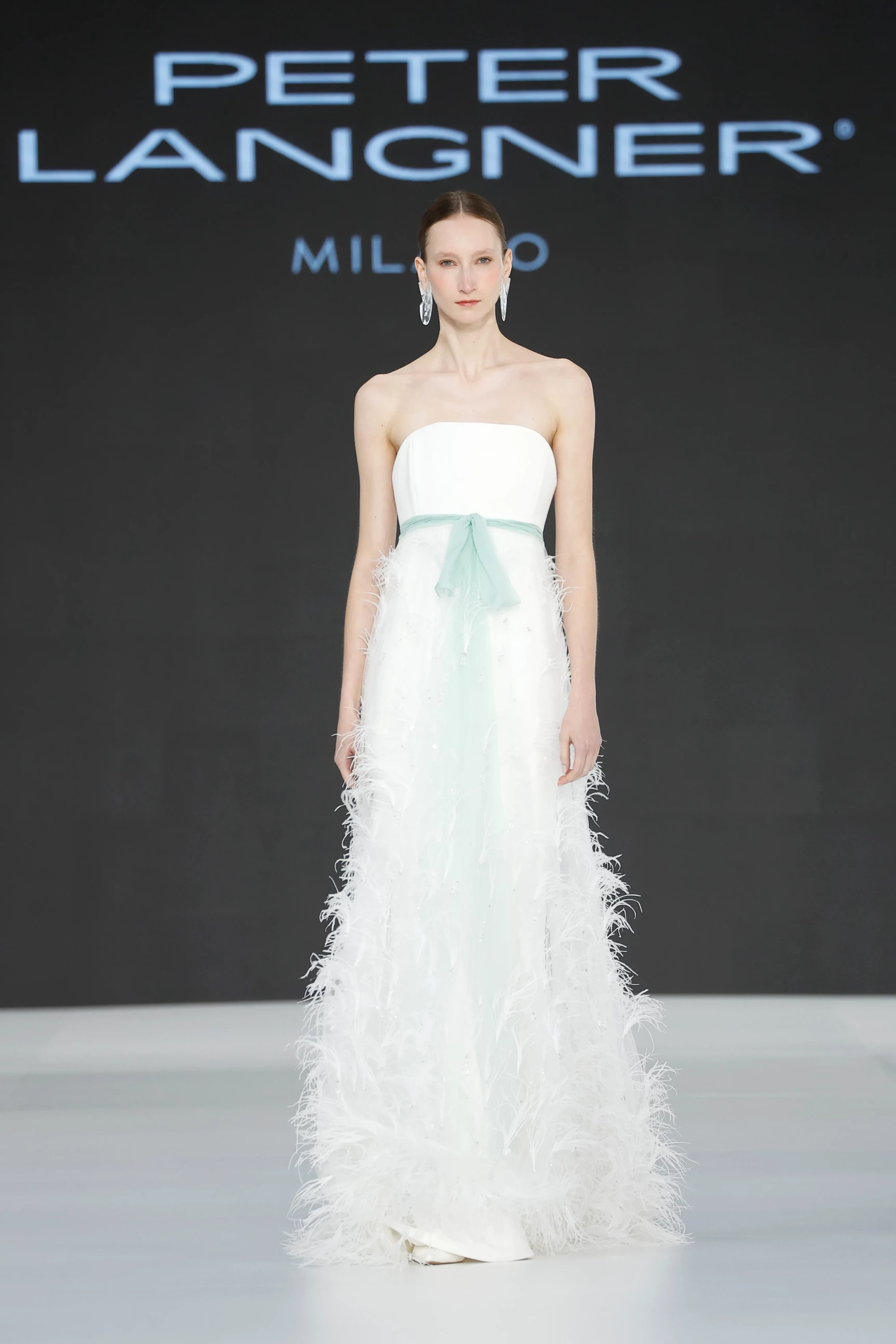 Barcelona Bridal Fashion Week 2026'da Peter Langner'ın 2027 İlkbahar gelinlik koleksiyonundan, pudra yeşili bir kuşakla vurgulanmış, straplez beyaz korsajlı ve eteği zarif tüylerle hareketlendirilmiş akışkan bir gelinlik modeli podyumda sergileniyor.