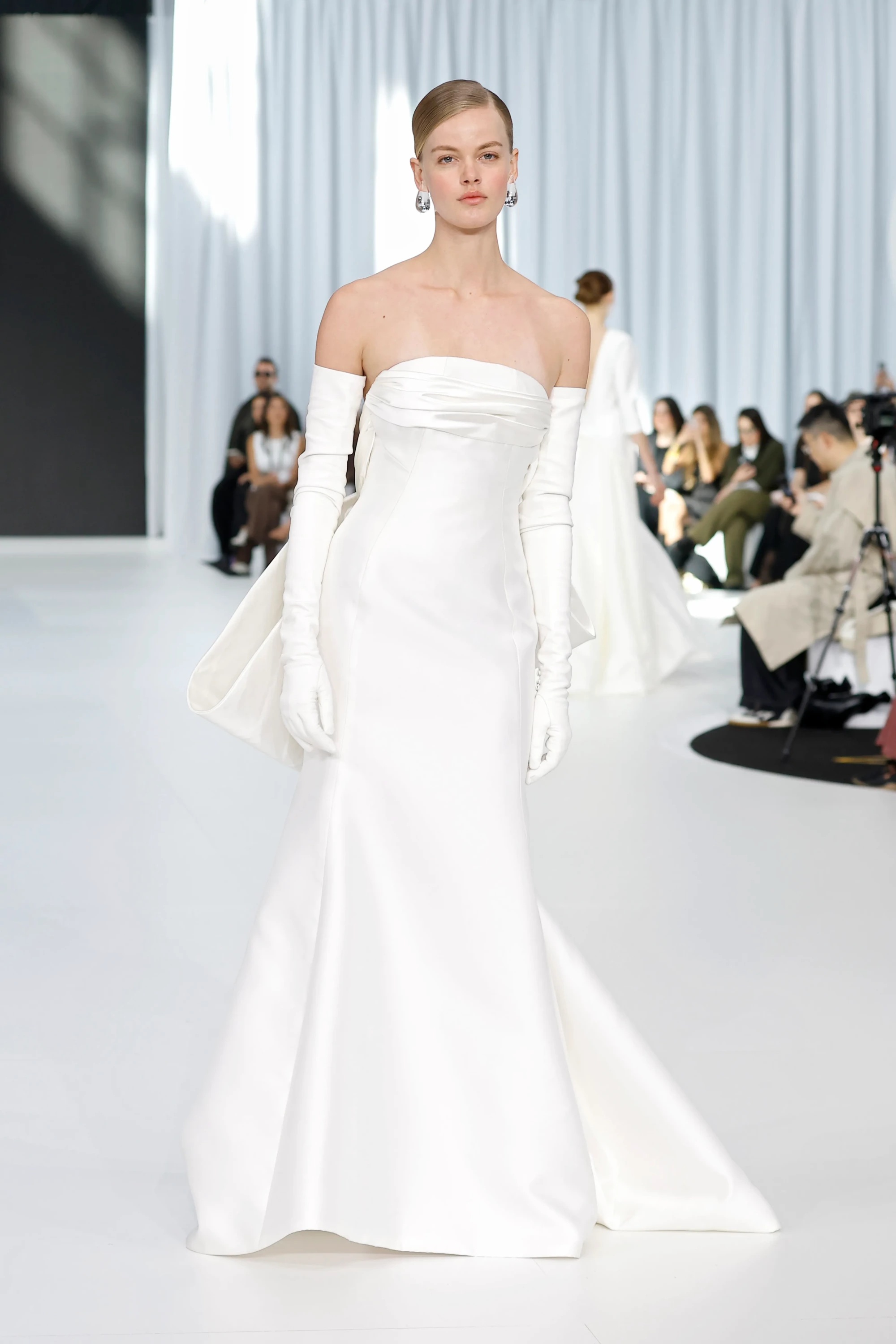 Barcelona Bridal Fashion Week 2026 defilesinde podyumda yürüyen model, straplez, zarif ve parlak saten kumaştan tasarlanmış, balık etek kesimli, uzun kuyruklu beyaz bir gelinlik giyiyor; görünümünü dirsek hizasındaki uzun beyaz eldivenler ve büyük damla küpelerle tamamlayarak 2027 gelinlik trendlerinden modern ve sofistike bir yorum sunuyor.