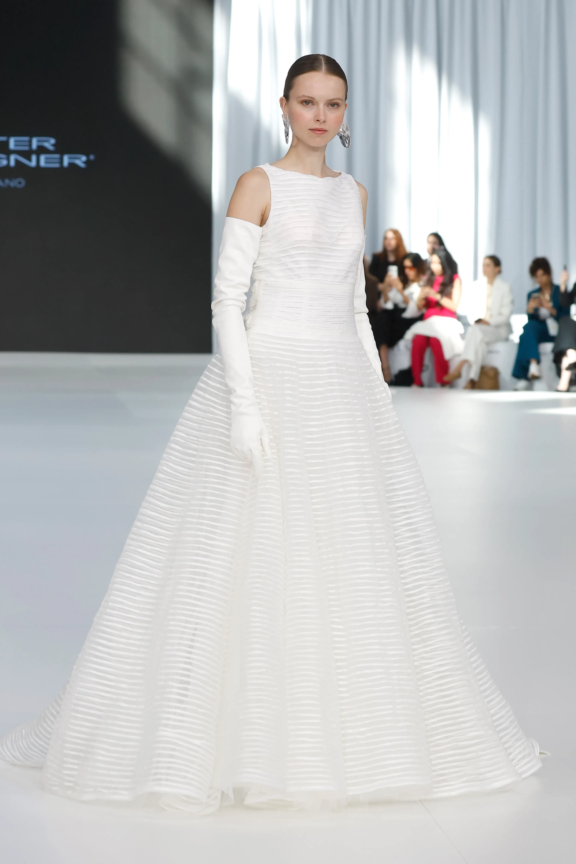 Peter Langner'in 2027 gelinlik trendlerini sergilediği Barcelona Bridal Fashion Week defilesinden, zarif bir topuzla saçları toplanmış, büyük şeffaf küpeler ve dirsek boyu eldivenlerle tamamladığı, yatay ribana dokulu, hacimli etekli ve kayık yakalı beyaz gelinliğiyle podyumda yürüyen bir model.