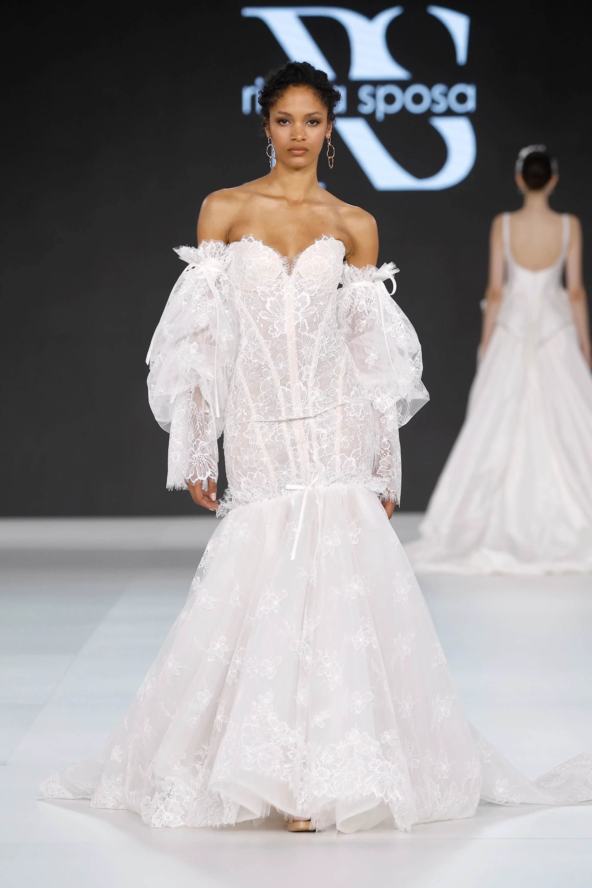 Siyah fonda, Ricca Sposa markasının adının önünde duran, bej tenli bir model, korse etkili, omuzları açık, üç boyutlu çiçek desenli dantellerle işlenmiş, hacimli kollara ve balık etek kesimine sahip beyaz bir gelinlik giyiyor; bu tasarım 2027 gelinlik trendlerini yansıtan feminen ve zamansız bir görünüm sunuyor.