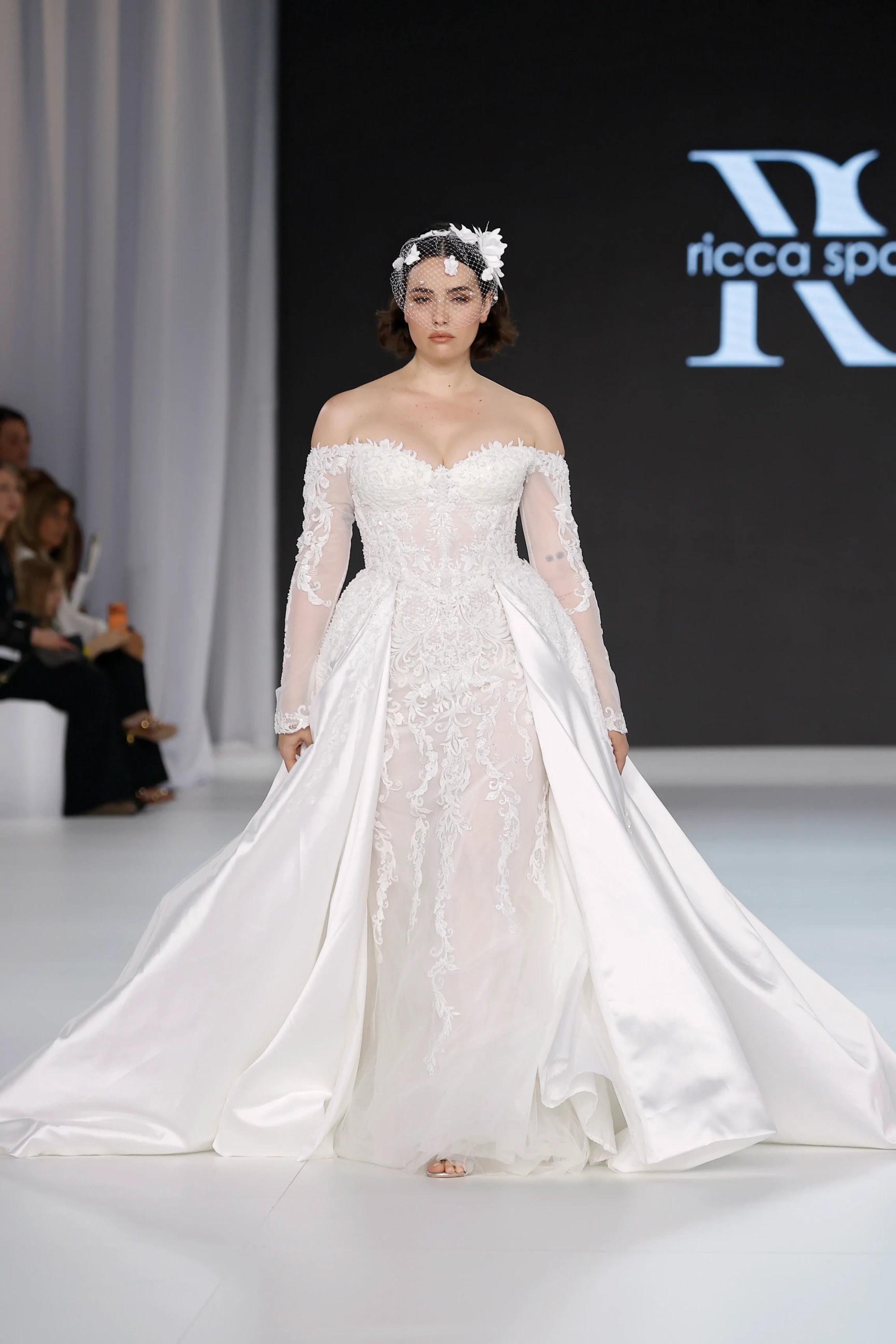 Beyaz bir podyumda, Ricca Sposa logosunun önünde yürüyen, fildişi rengi, omuzları açık, şeffaf dantel kollara ve işlemeli dar bir üst bedene sahip, önü açık kabarık saten etekli görkemli bir gelinlik giymiş model. Bu tasarım, 2027 Gelinlik Trendleri arasında romantik bir vurgu yaparak, zarif detayları ve akışkan siluetiyle dikkat çekiyor. Modelin yüzünü kapatan çiçek ve inci süslemeli kısa tül duvağı ise modern ve sofistike bir dokunuş katıyor.