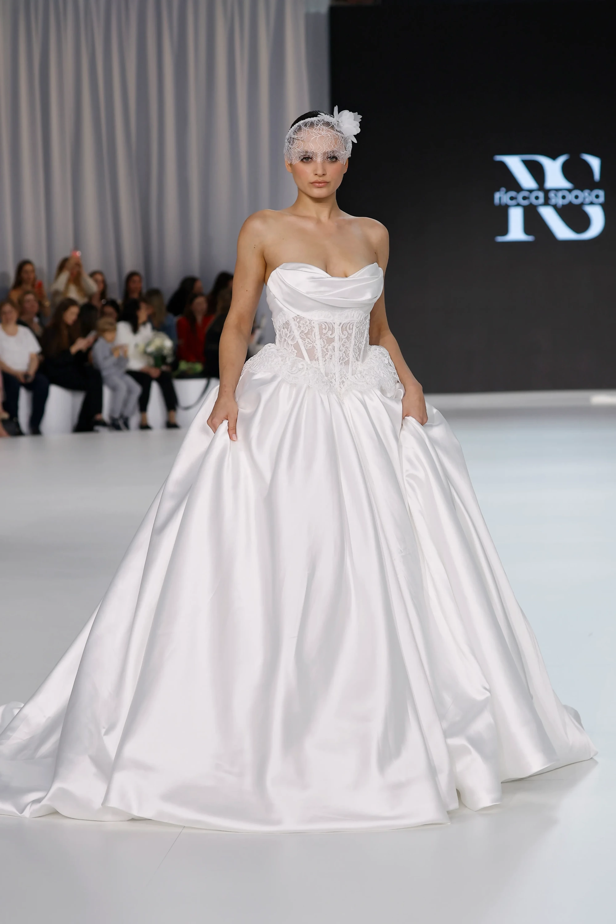 Podyumda yürüyen bir model, Ricca Sposa'nın 2027 gelinlik trendlerinden ilham alan, straplez kalp yaka dantel korsajı ve hacimli saten etekleriyle feminen silüeti vurgulayan modern romantik beyaz bir gelinlik giyiyor, alnını zarifçe örten çiçek detaylı dantel bir duvak tamamlıyor.