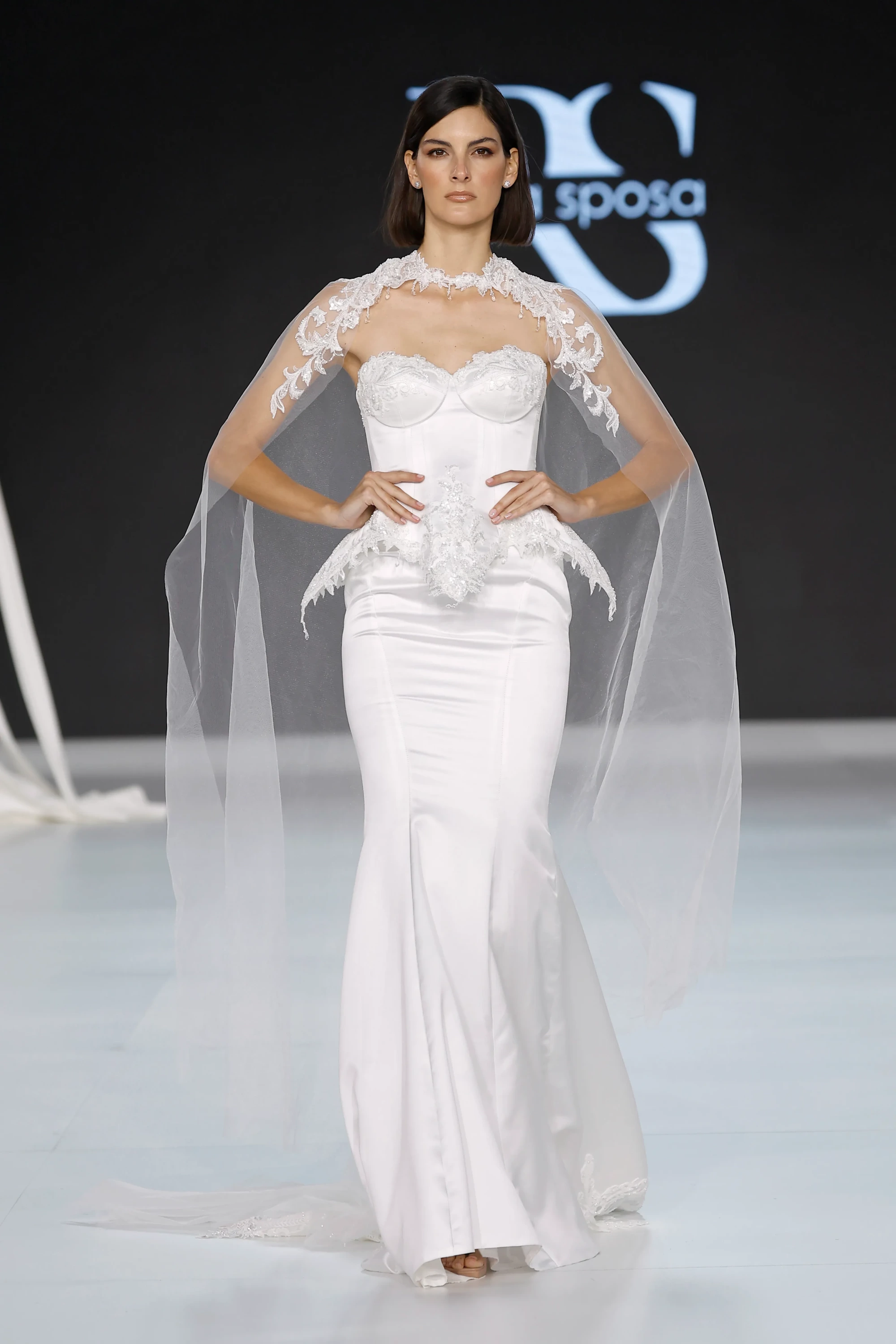 Barcelona Bridal Fashion Week 2026'da sergilenen Ricca Sposa İlkbahar 2027 gelinlik koleksiyonundan, vücuda oturan beyaz dantel elbisesi, kalp yaka korsajı, belindeki volanlı detayları ve omuzlardan sarkan transparan, zarif dantel işlemeli pelerini ile modern romantizmi yansıtan bir model podyumda duruyor.