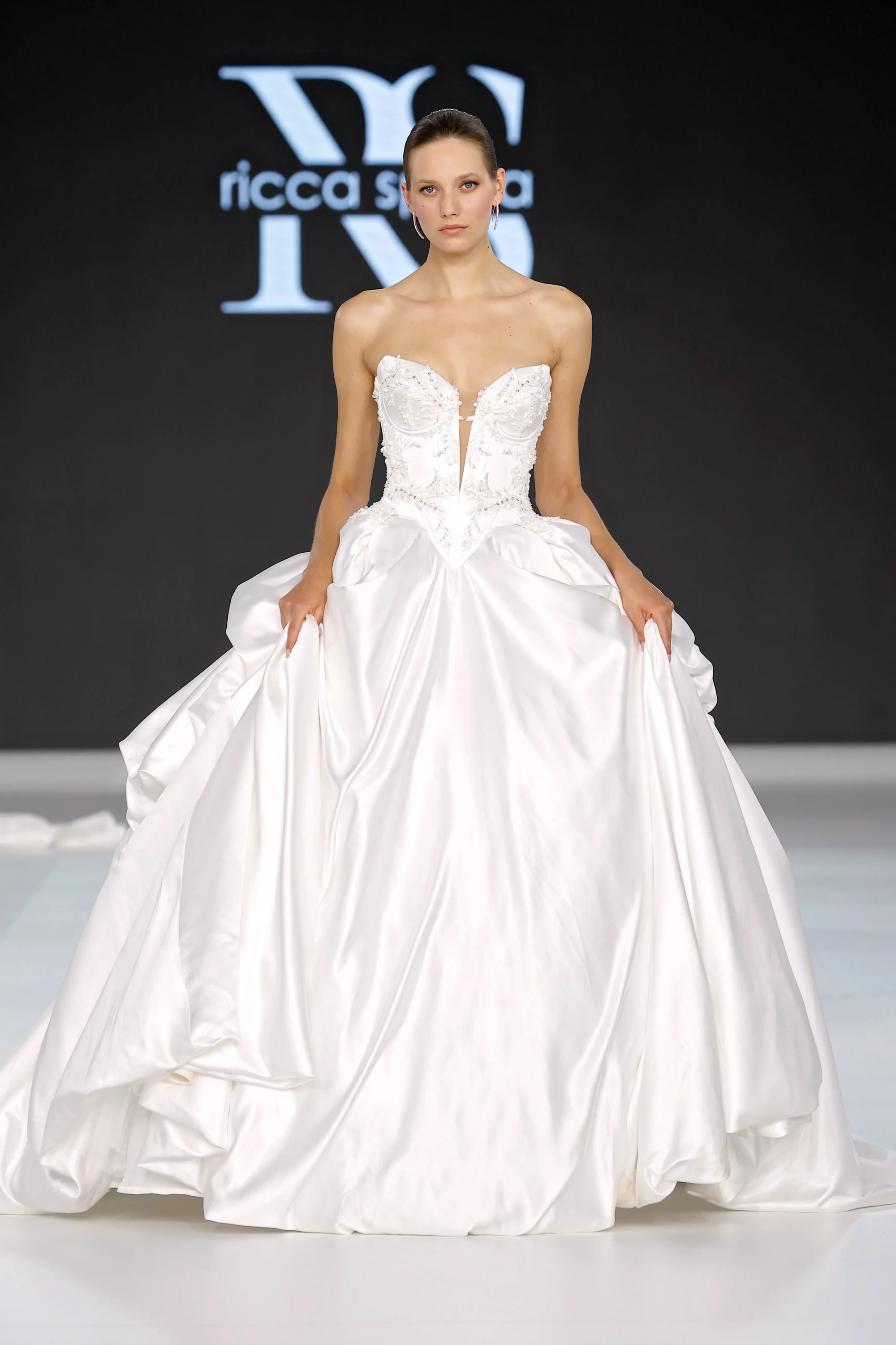 Model, Ricca Sposa & Yuliia Lobachóva Couture İlkbahar 2027 koleksiyonundan, zengin işlemeli kalp yaka korsajı ve kabarık saten eteğiyle dikkat çeken, 2027 gelinlik trendlerini yansıtan beyaz, straplez bir prenses kesim gelinlik giyiyor.