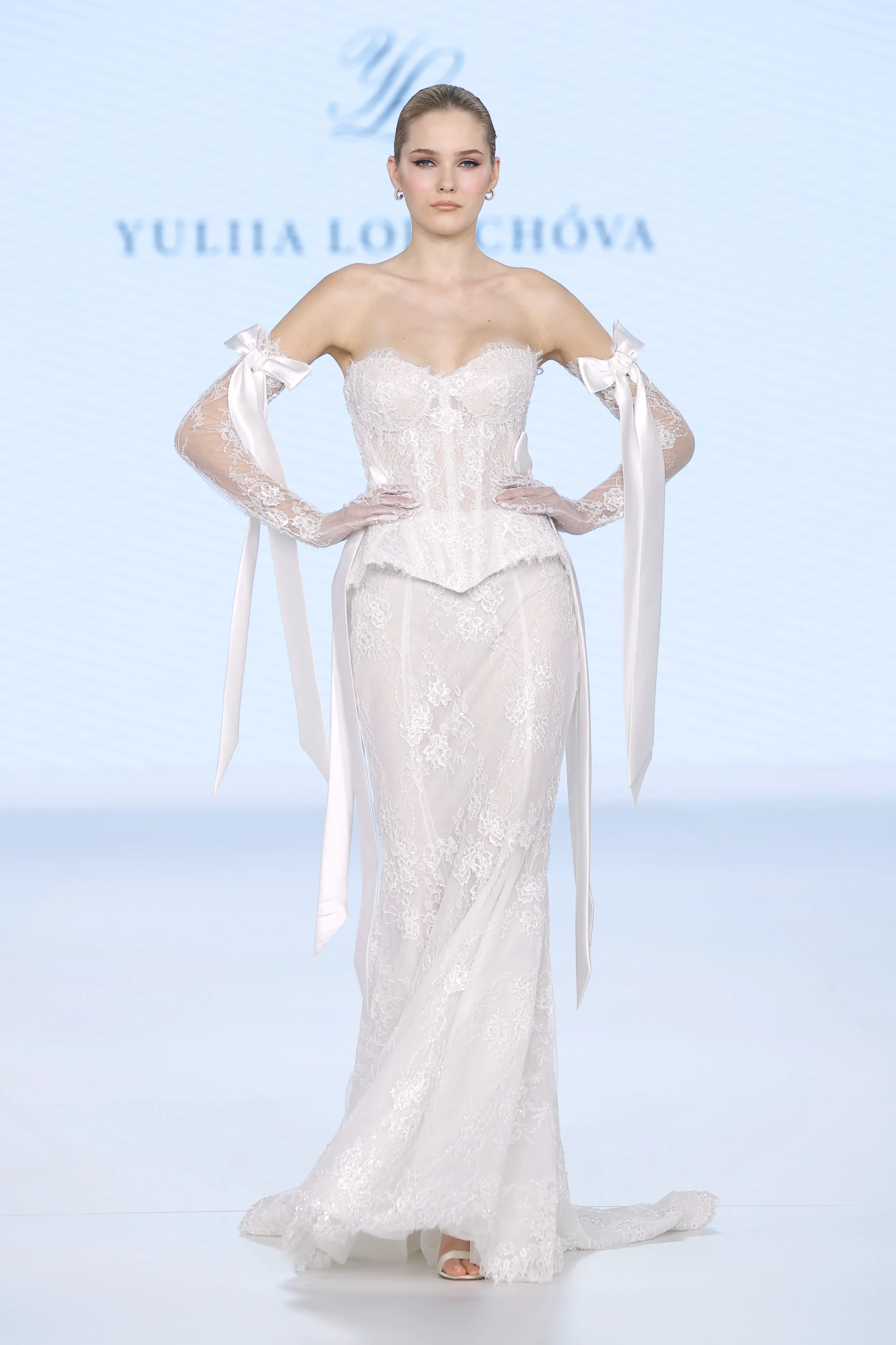 YULIIA LOBACHOVA Couture İlkbahar 2027 gelinlik koleksiyonundan, podyumda elleri belinde poz veren, modern ve romantik bir gelinlik giyen model; kalp yaka korsajlı, vücuda oturan dantel elbisesini, kollarına bağladığı uzun saten kurdele detaylı transparan dantel eldivenlerle tamamlayarak 2027 gelinlik trendlerini yansıtıyor.