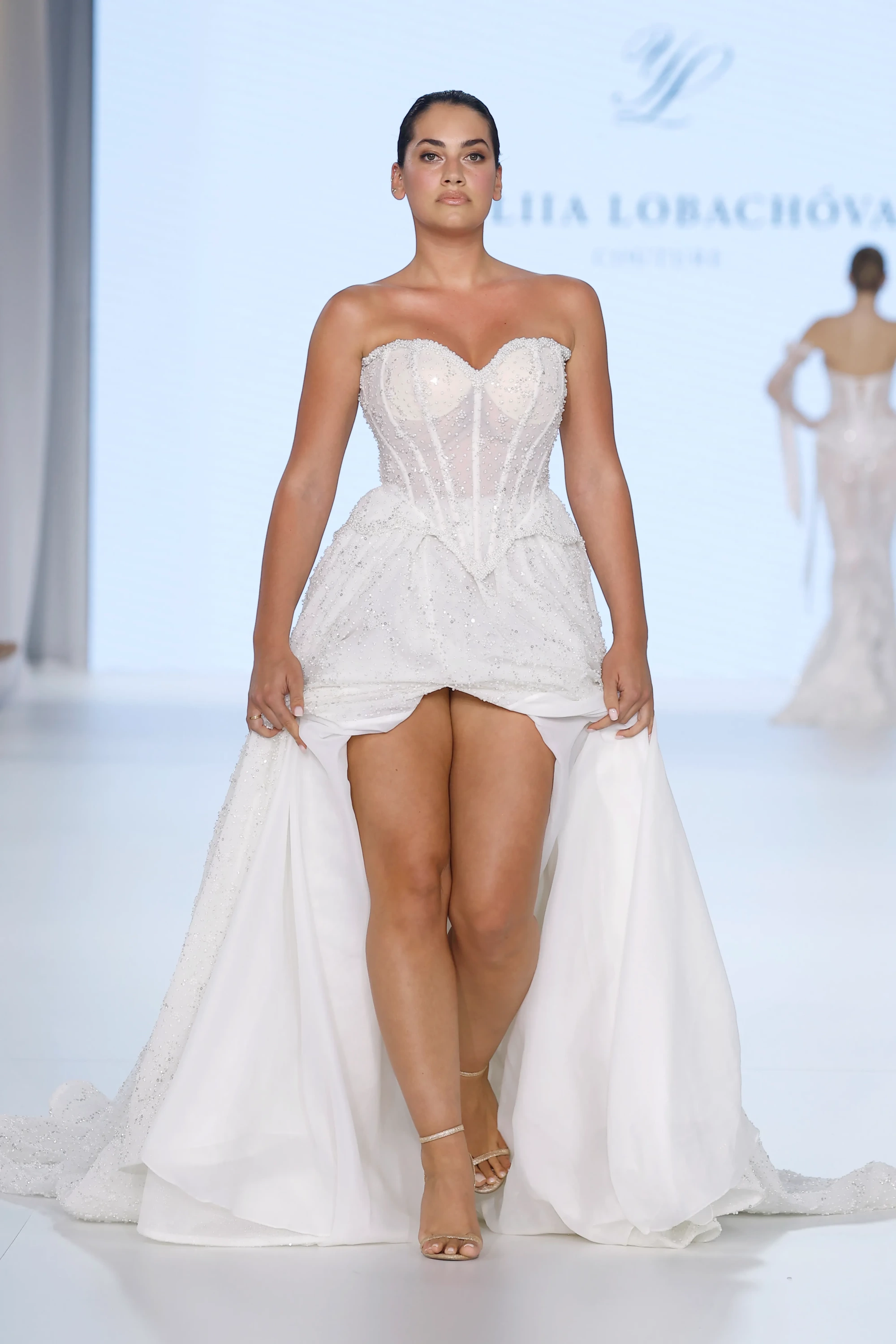 Podyumda yürüyen bir model, Ricca Sposa & Yuliia Lobachóva Couture'dan, transparan detaylı, işlemeli kalp yaka korsajı ve ışıltılı, akışkan eteğini kaldırarak bacaklarını sergilediği modern ve romantik bir 2027 gelinlik trendini cesurca sunuyor.