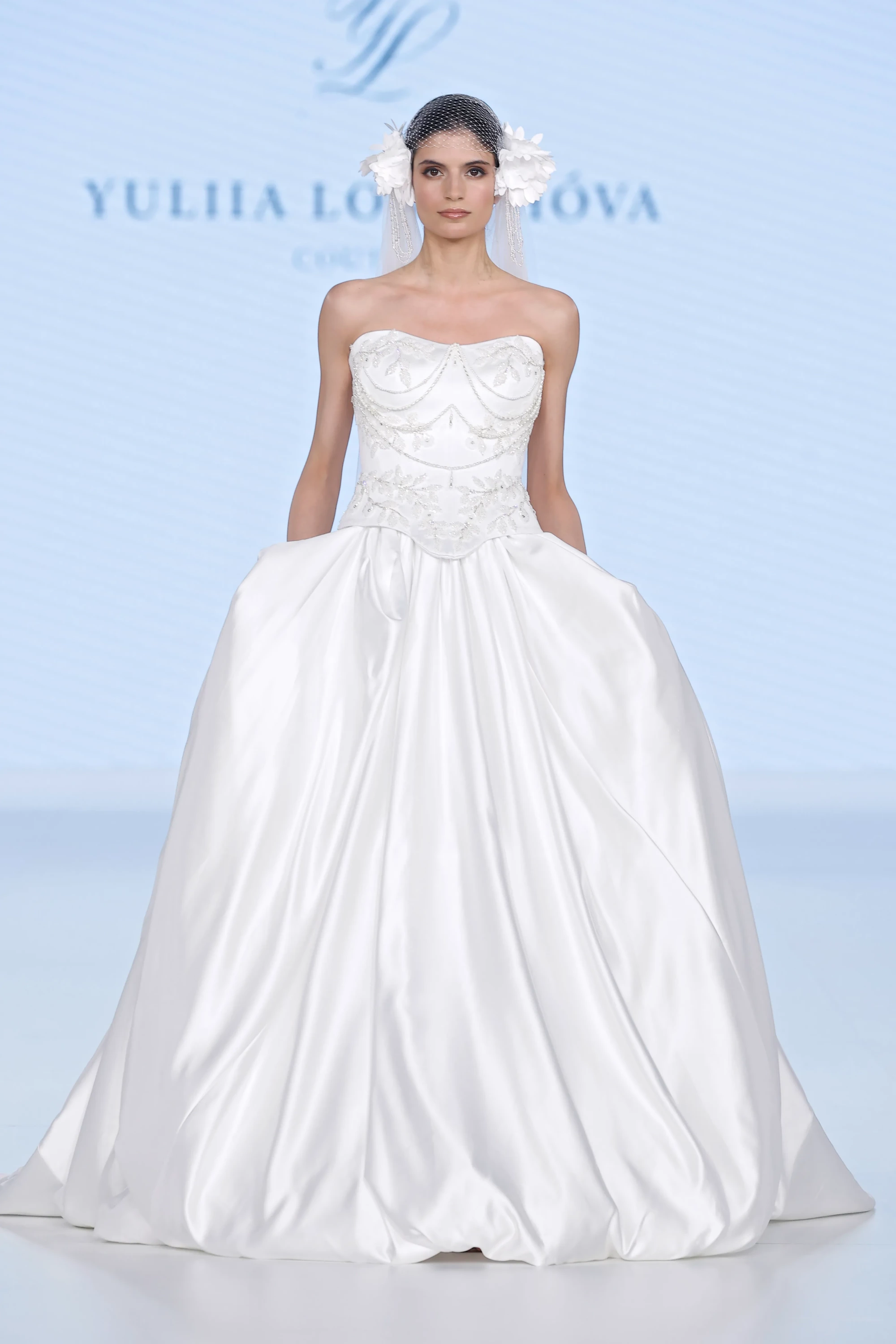 Barcelona Bridal Fashion Week 2026 podyumunda, Ricca Sposa & Yuliia Lobachova Couture İlkbahar 2027 gelinlik trendlerinden modern ve romantik bir tasarımı tanıtan model; straplez işlemeli korsajı, kabarık saten eteği ve file duvak üzerine yerleştirilmiş 3D çiçeklerle süslü saç aksesuarlarıyla oldukça göz alıcı duruyor.