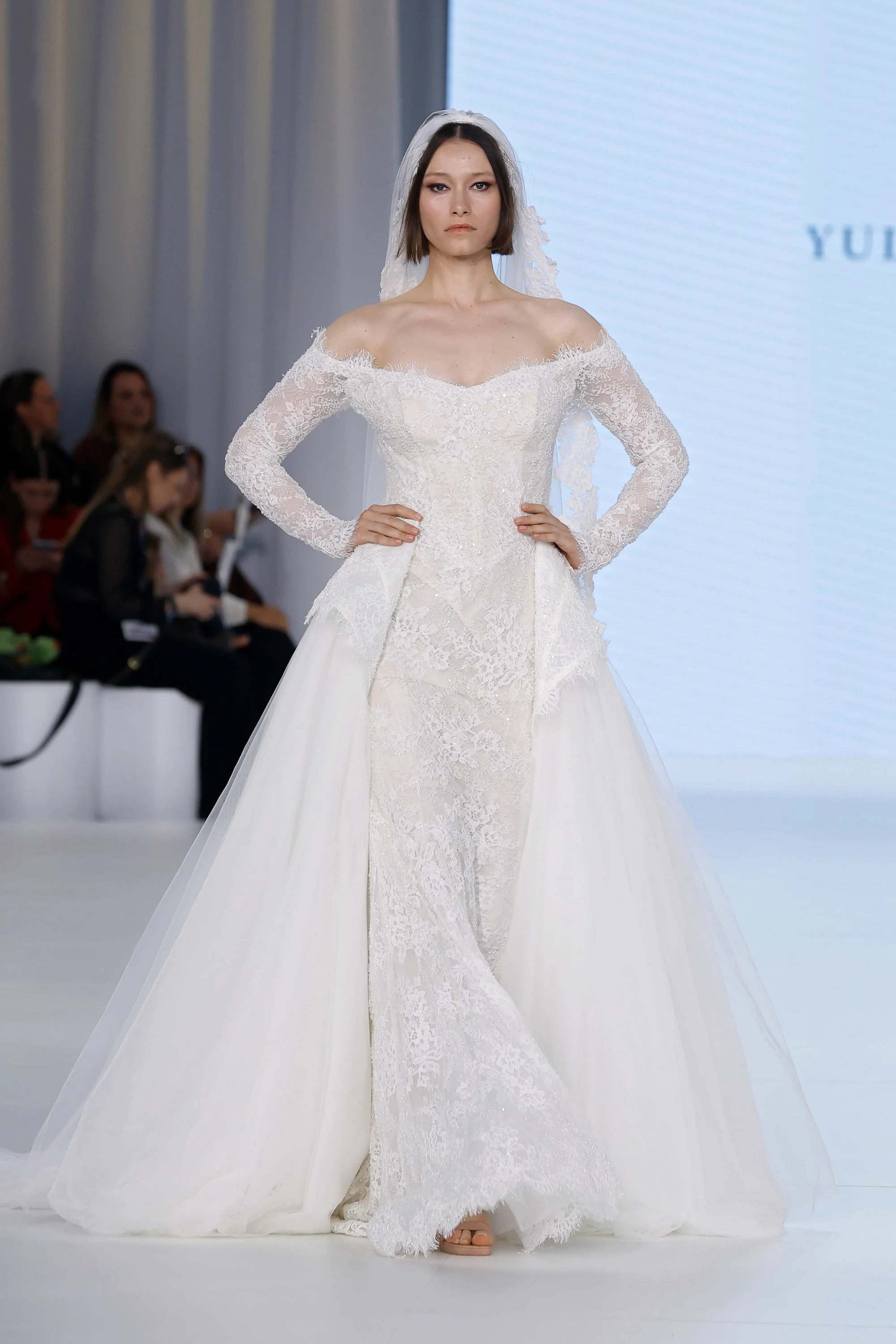 Model, 2027 gelinlik trendlerini yansıtan, Ricca Sposa Yuliia Lobachóva Couture markalı, omuzları açık kalp yakalı, transparan dantel kollara ve parıltılı vücuda oturan dantel korsaja sahip, kabarık tül dış etekle dramatik bir kuyruk oluşturan modern bir gelinliği podyumda sergiliyor.