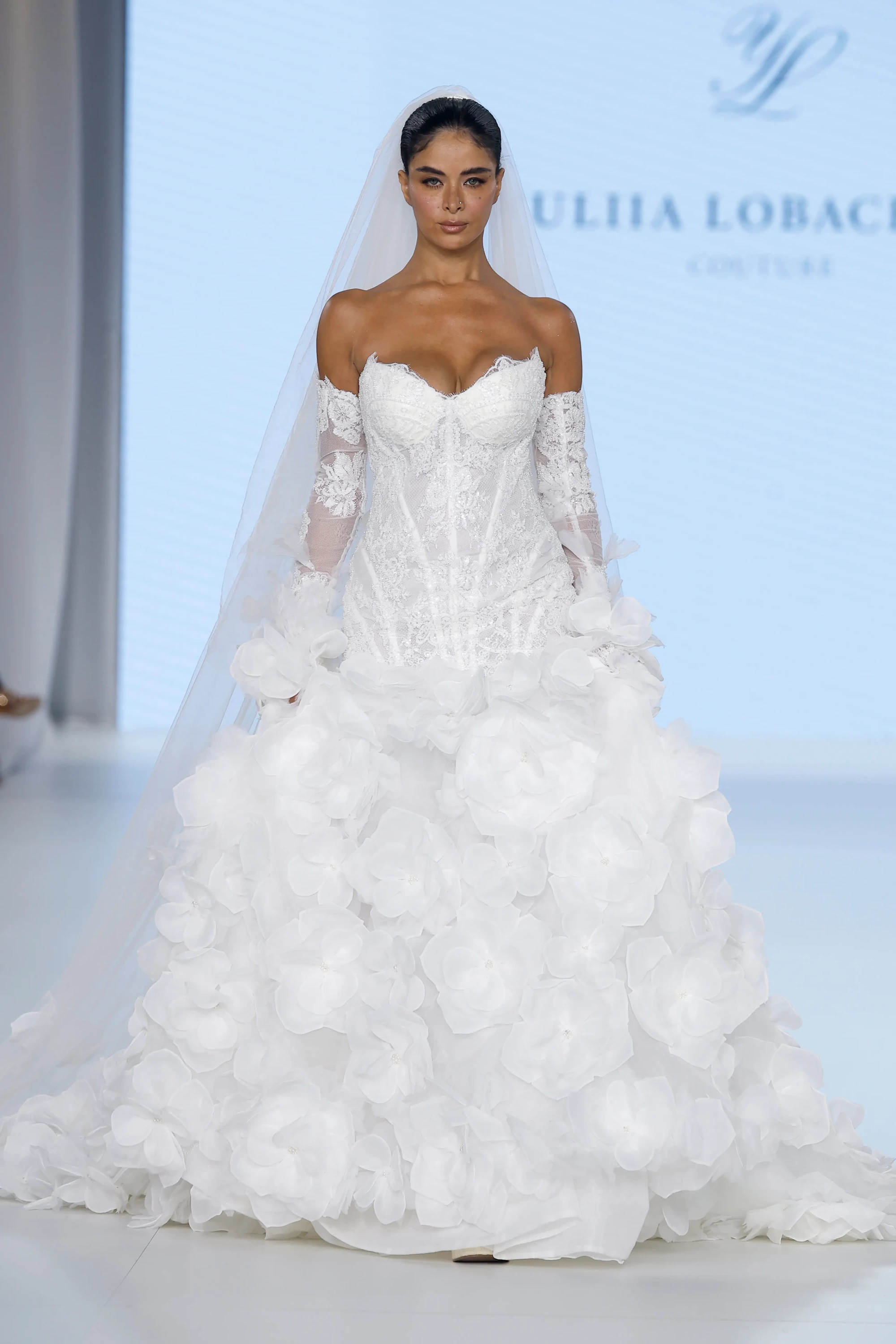 Podyumda, Ricca Sposa & Yuliia Lobachóva Couture imzalı, askısız dantel korsajlı üstü, şeffaf uzun kolları fırfır detaylı ve hacimli eteği büyük üç boyutlu çiçek aplikeleriyle kaplı modern bir gelinlik giymiş model, 2027 gelinlik trendlerinden romantik bir görünüm sergiliyor.