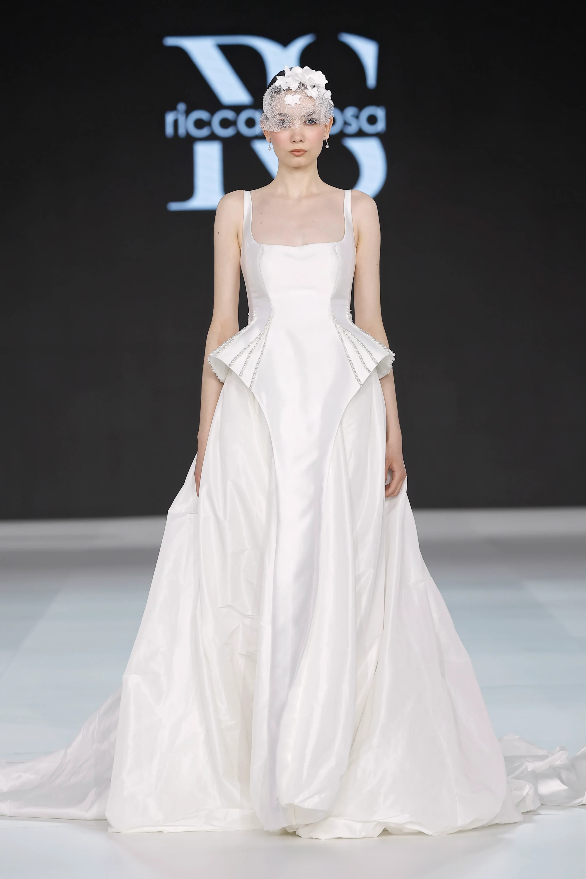 Bir model, Ricca Sposa'nın podyumunda, inci detaylı peplum korsajı ve hacimli eteğiyle modern bir yorum kazanan beyaz gelinliğini sergiliyor; başındaki üç boyutlu çiçek aplikeli tül duvak, 2027 gelinlik trendlerinden romantik ve yenilikçi bir görünüm sunuyor.