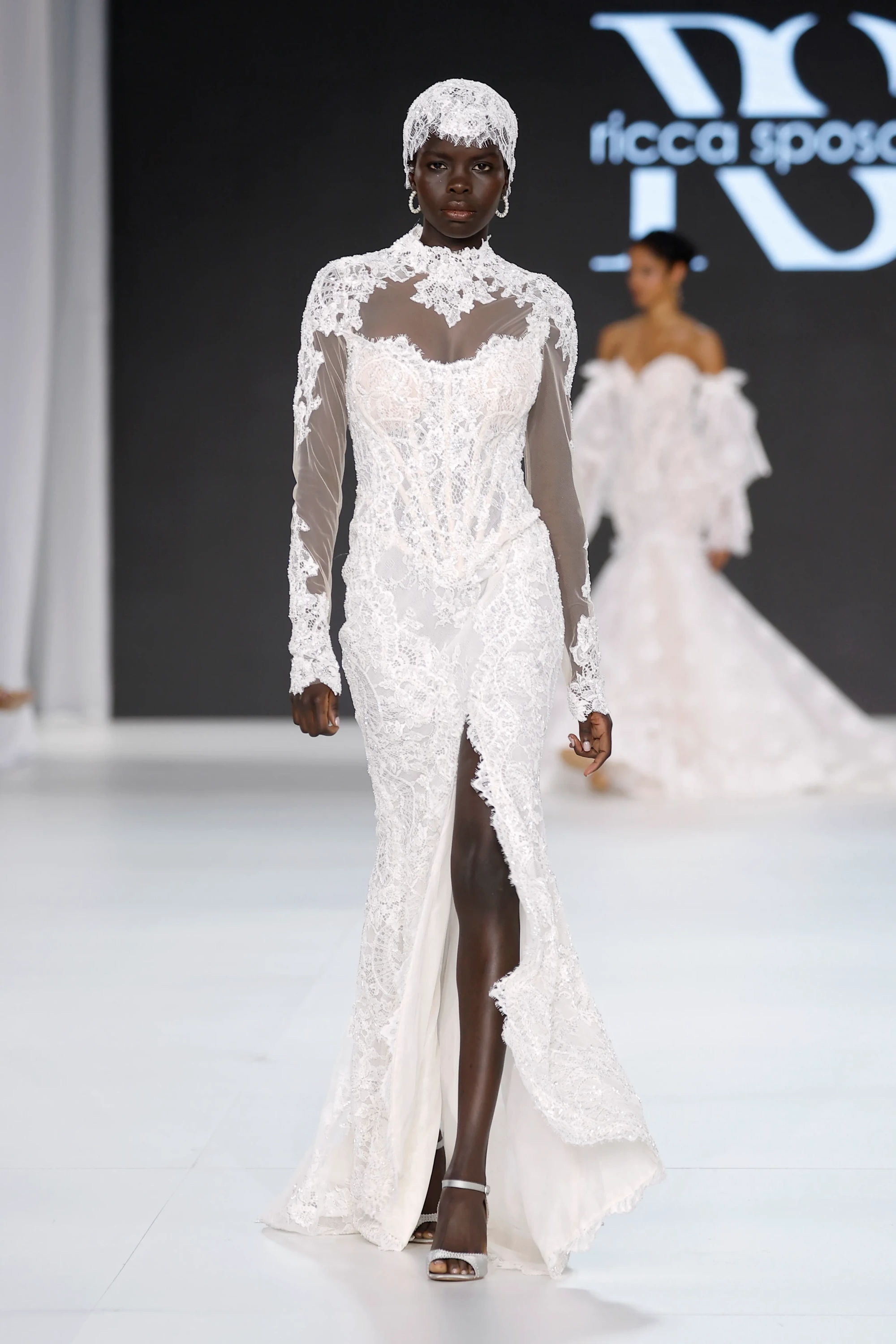 Ricca Sposa & Yuliia Lobachóva Couture İlkbahar 2027 Gelinlik Koleksiyonu'ndan, dantel başlığı, transparan detaylı yüksek yakası, uzun kolları ve bacak yırtmacıyla öne çıkan, incelikli dantel işlemeli bir gelinlik, 2027 gelinlik trendlerinde romantik ve feminen bir çizgiyi sergiliyor.