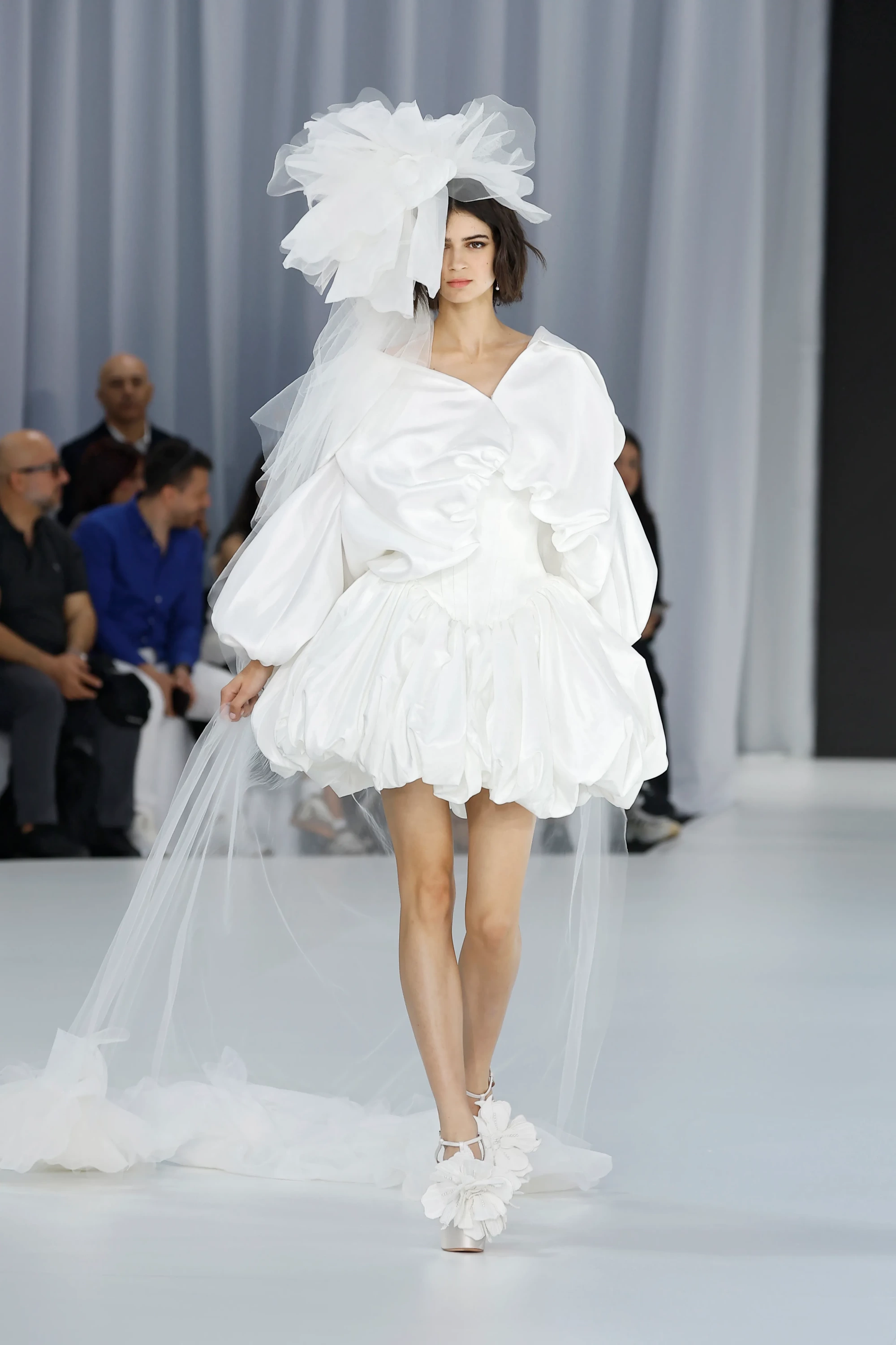 Barcelona Bridal Fashion Week'te Serina'nın 2027 İlkbahar gelinlik koleksiyonundan, kabarık kısa bir elbise, yüzünü kısmen kapatan büyük, heykelsi bir tül başlık, uzun şeffaf duvak ve 3D çiçek aplikeli platform ayakkabılar giymiş bir model podyumda ilerliyor.