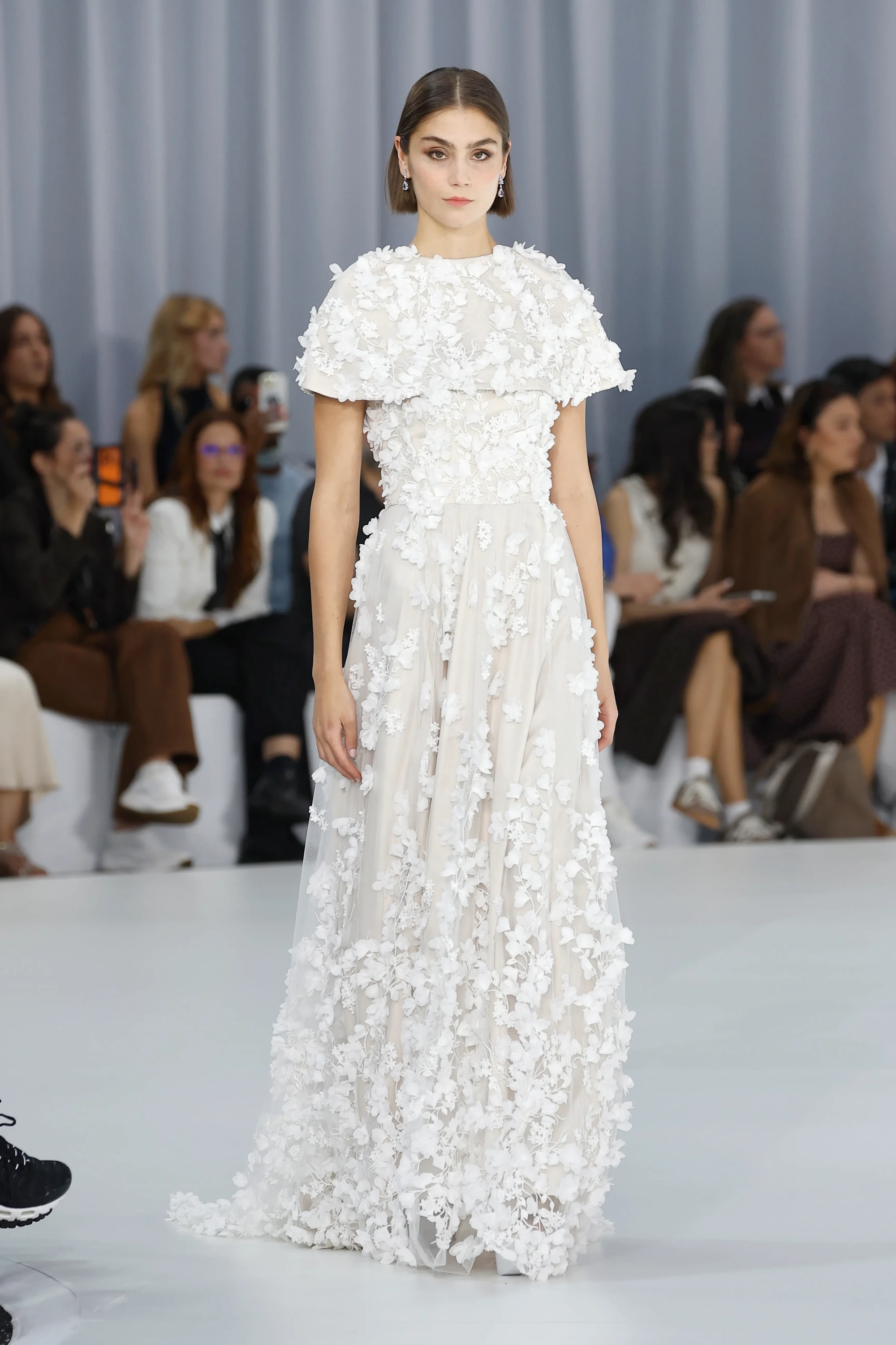 Barcelona Bridal Fashion Week 2027 İlkbahar koleksiyonundan Serina markasının podyumunda yürüyen kısa saçlı model, ekru astarlı şeffaf tül üzerine yoğun bir şekilde serpiştirilmiş, üç boyutlu beyaz çiçek aplikeleriyle süslü ve omuzları saran katmanlı çiçekli pelerin detayıyla doğanın akışkan romantizmini yansıtan bir gelinlik sergiliyor.