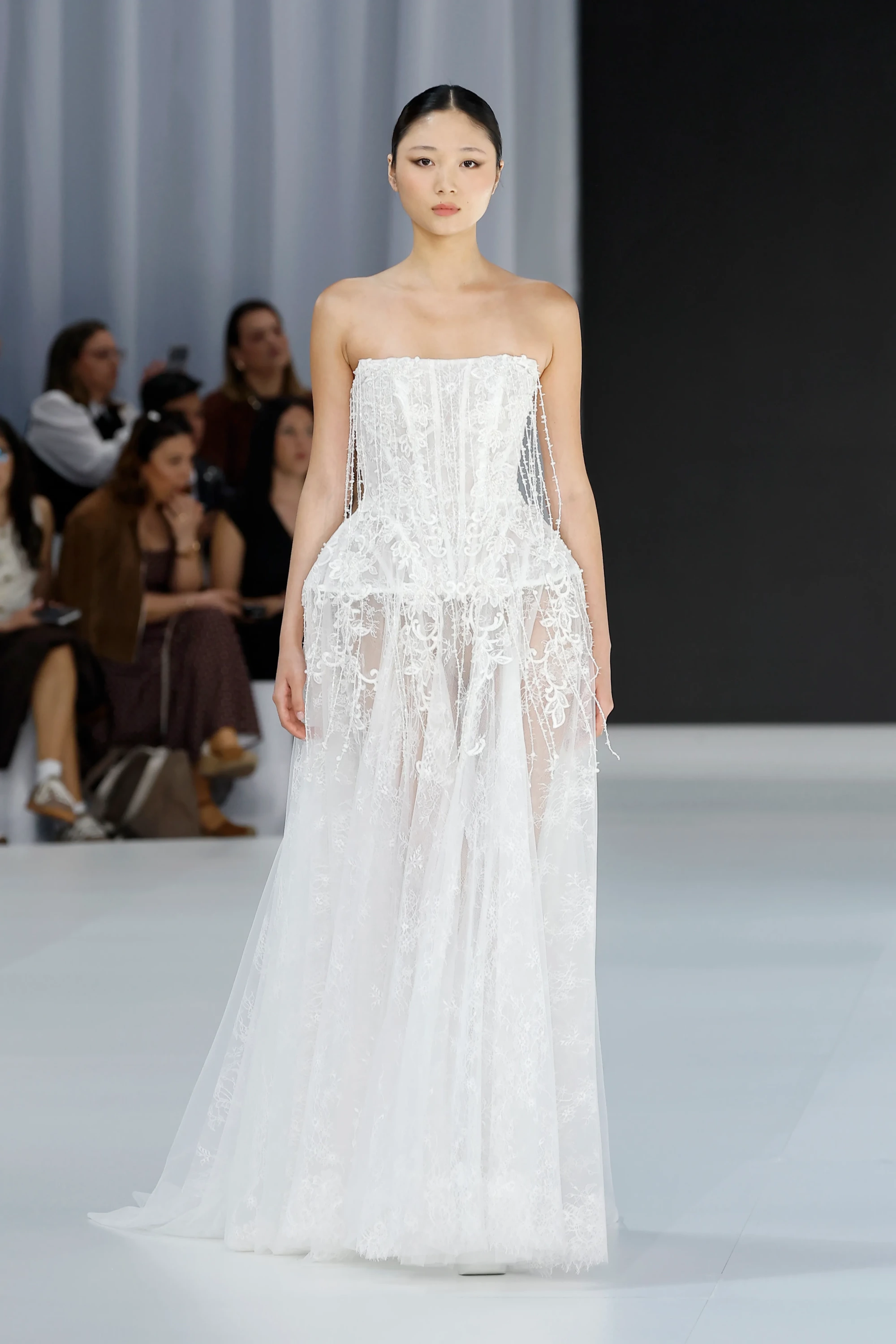 Barcelona Bridal Fashion Week'te Serina'nın İlkbahar 2027 gelinlik koleksiyonundan, podyumda duran zarif model, inci işlemelerle süslenmiş straplez korsajı ve transparan tül katmanları ile akışkan bir siluet oluşturan, doğadan ilham alan çiçek aplikeleriyle bezenmiş romantik beyaz bir gelinlik sergiliyor.