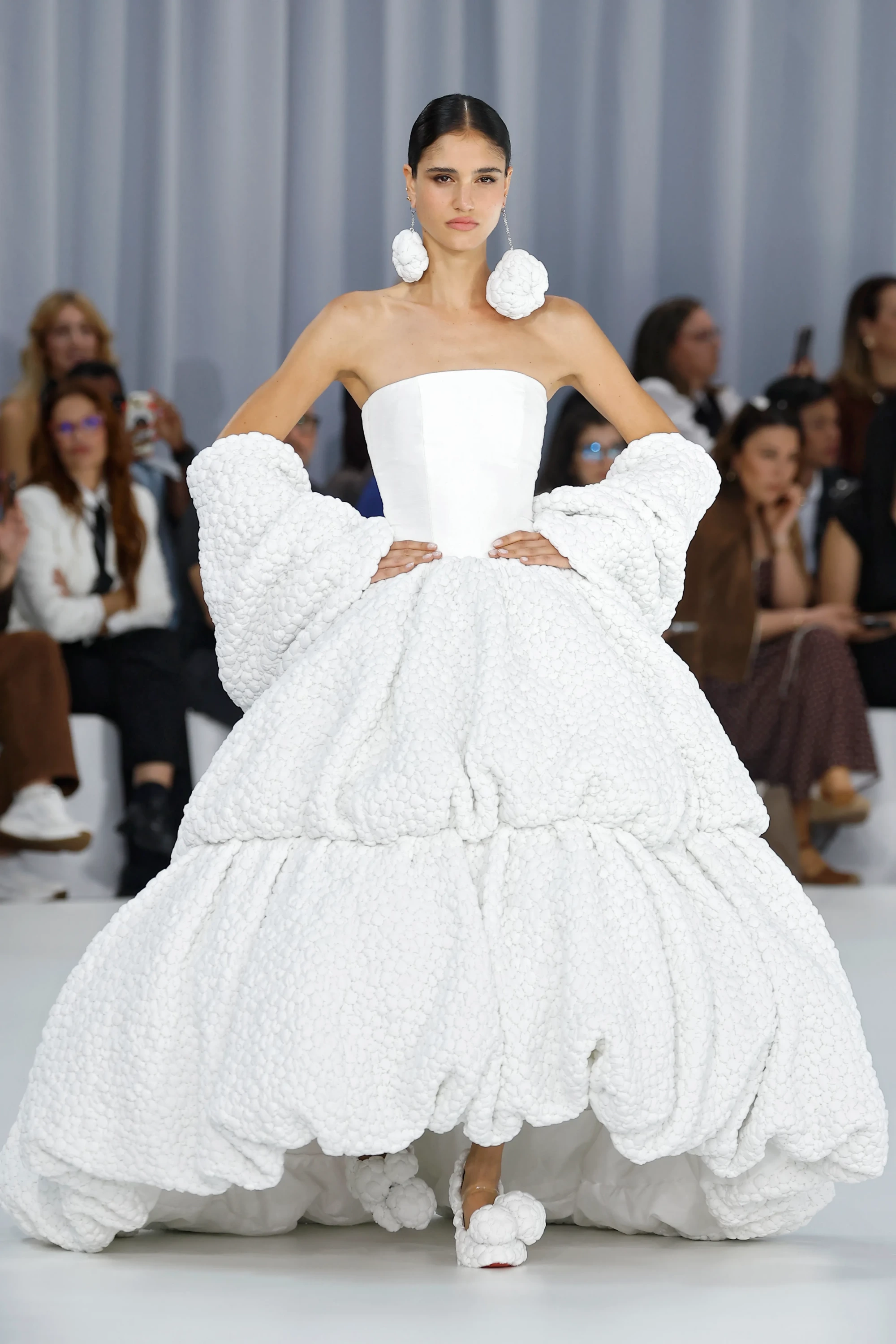 Barcelona Bridal Fashion Week podyumunda, straplez, kabarık ve katmanlı, kabartmalı dokulu beyaz bir gelinlik giyen model, aynı dokuya sahip iri küpeler ve ayakkabılarla 2027 gelinlik trendleri arasında yer alan hacimli ve tekstürel tasarımları sergiliyor.