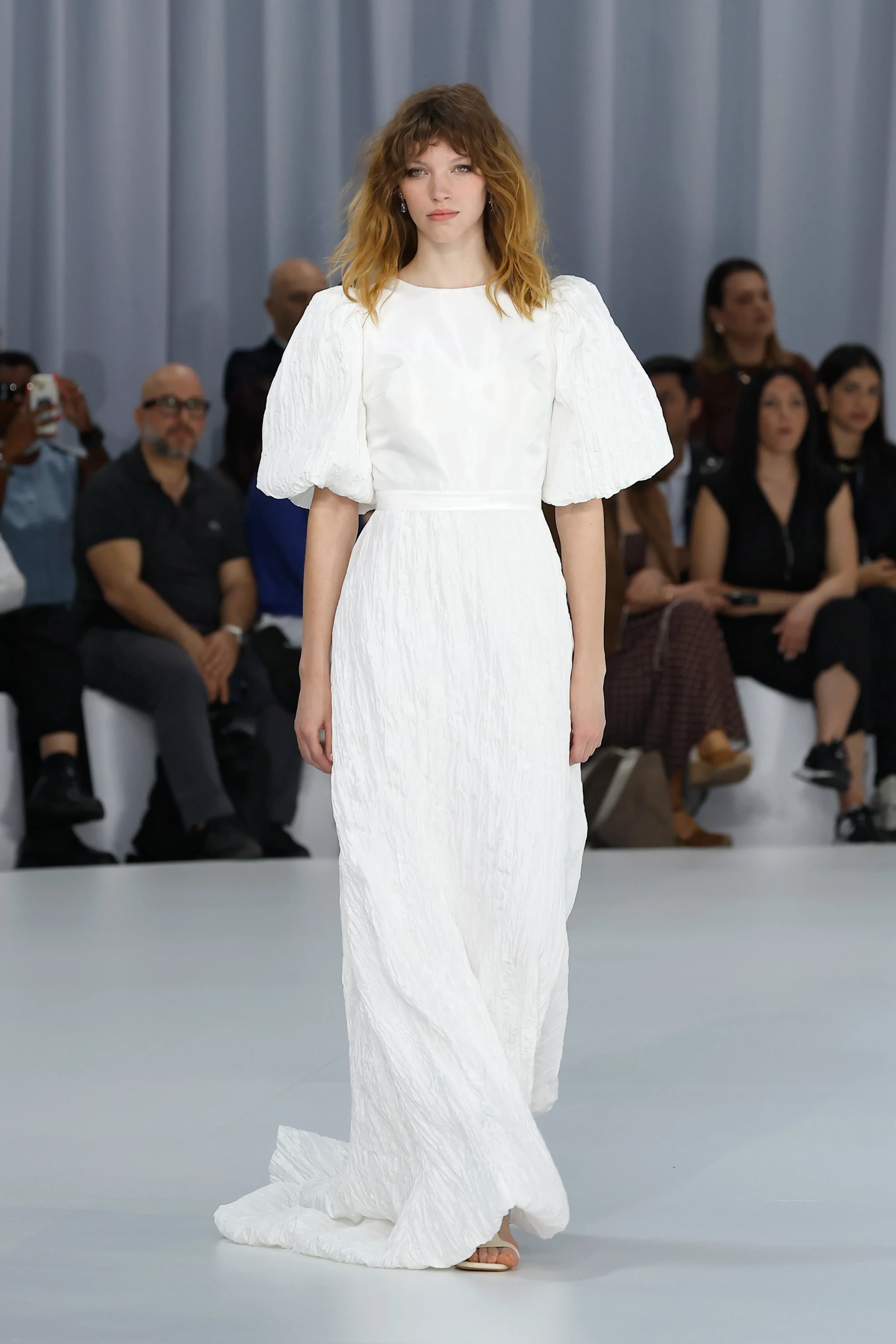 Barcelona Bridal Fashion Week kapsamında Serina'nın 2027 İlkbahar Gelinlik Koleksiyonu'ndan podyumda sunulan, dalgalı saçlı genç bir modelin giydiği, kabarık kısa kollu, bisiklet yaka, beli ince bir şeritle vurgulanmış ve hafif bürümcük dokulu, akışkan formlu, yere kadar uzanan bembeyaz gelinlik, modern romantizmi ve dinginliği temsil ediyor.