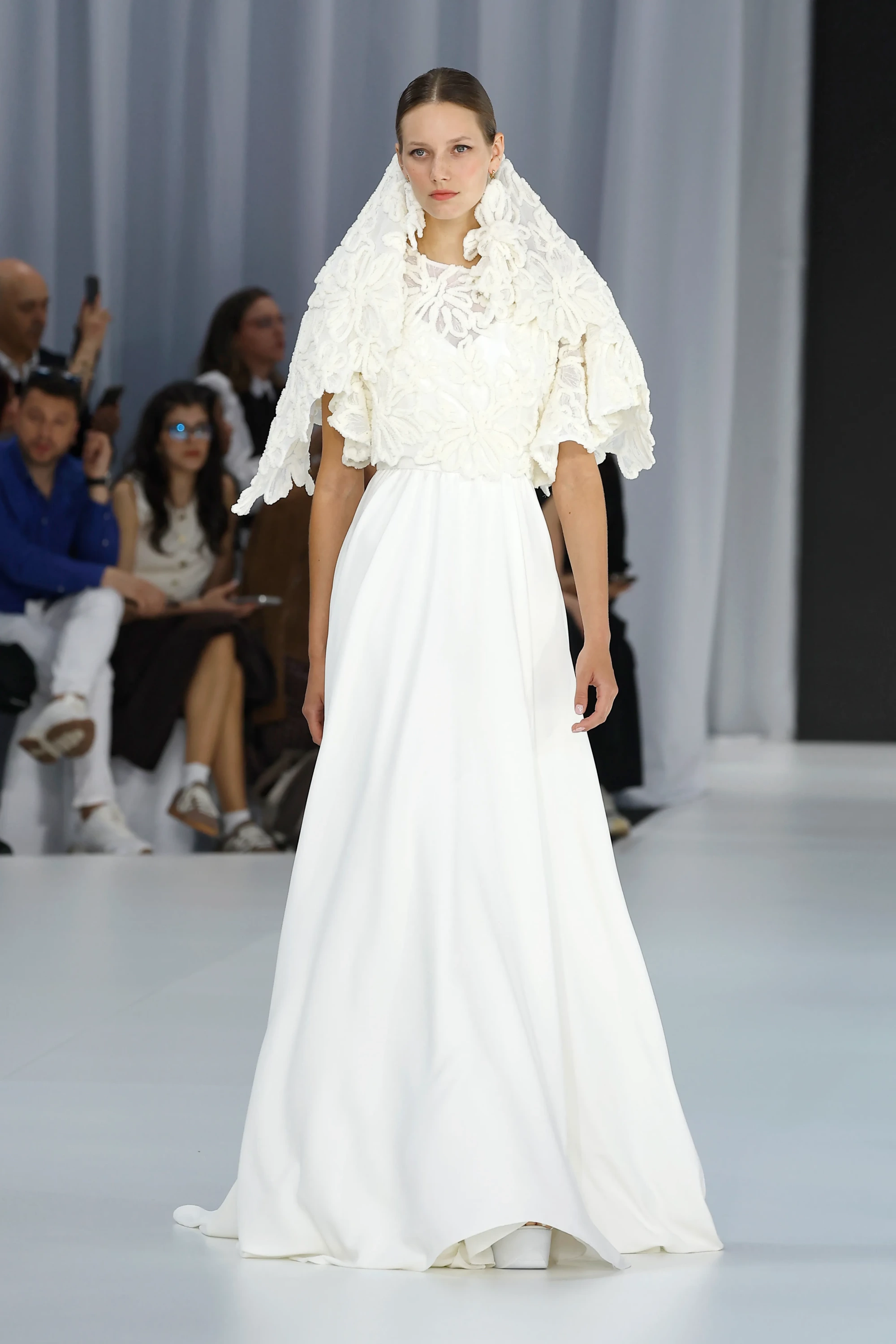Barcelona Bridal Fashion Week'te Serina'nın İlkbahar 2027 Gelinlik Koleksiyonu'ndan, organik detaylı ve kabartmalı çiçek motifleriyle zenginleştirilmiş volümlü bir yaka ve omuzlardan dökülen başlık ile alt kısmı sade ve akışkan formda bembeyaz bir gelinlik içinde yürüyen model, 2027 gelinlik trendlerinin romantik yorumunu gözler önüne seriyor.