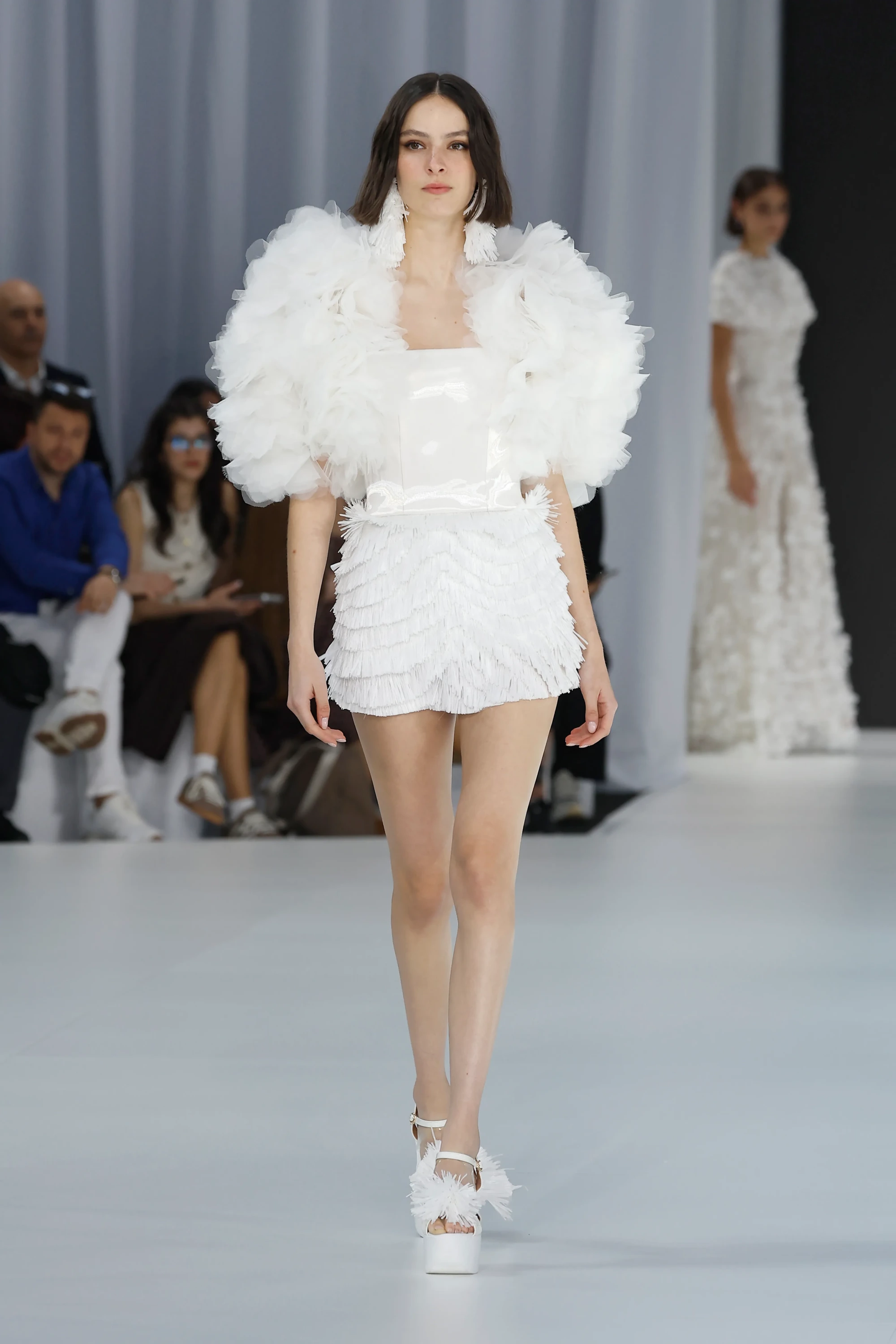 Serina İlkbahar 2027 Gelinlik Koleksiyonu'ndan, Barcelona Bridal Fashion Week podyumunda sergilenen, hacimli tül omuzları, katmanlı püsküllü mini eteği ve ponpon detaylı platform ayakkabılarıyla modern ve oyunbaz bir gelinlik trendi sergileyen model.