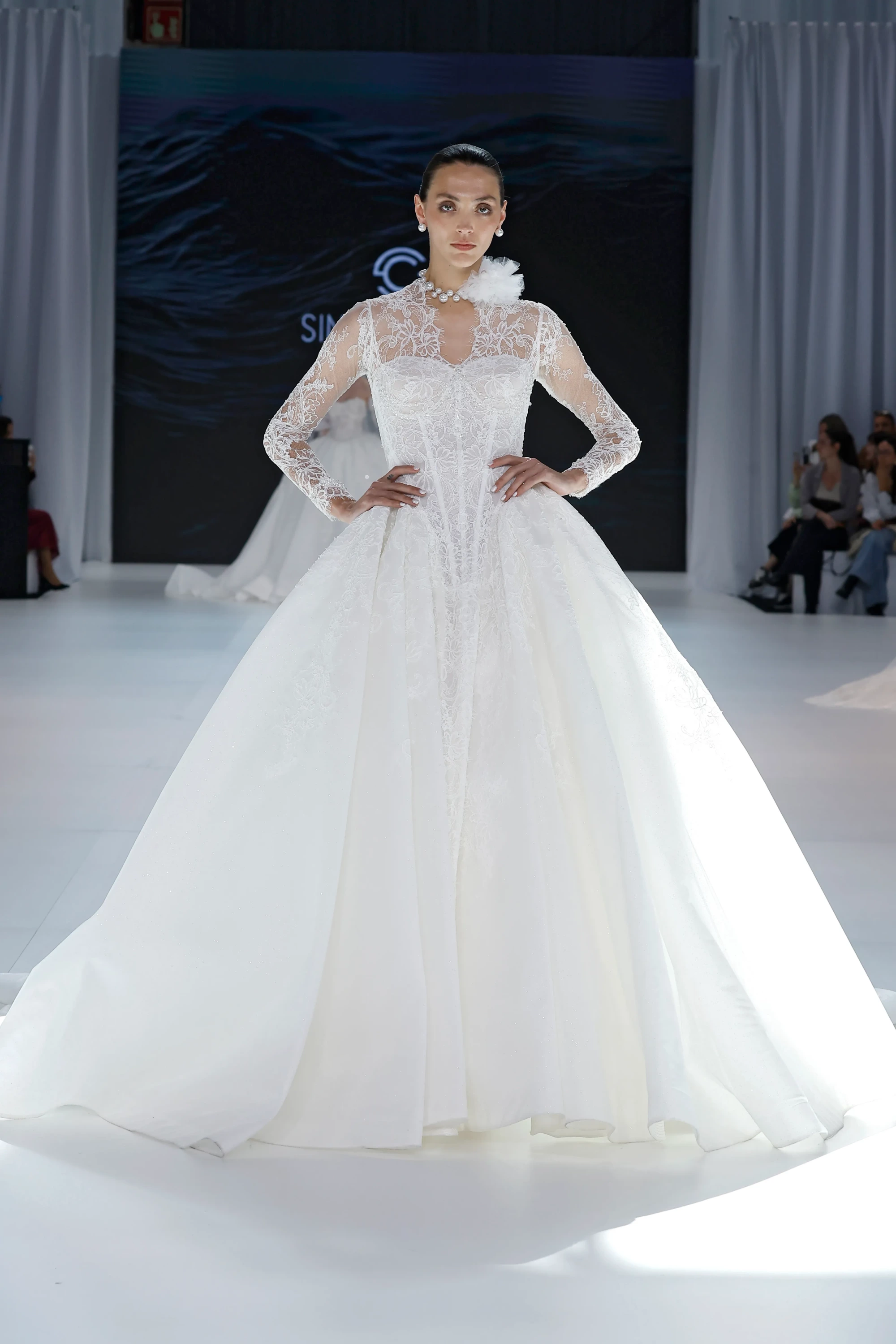 Podyumda duran bir model, kabarık etekli, zarif dantel uzun kollu ve korse etkili, bembeyaz bir Sima Couture gelinlik giymiş; inci kolye ve boynundaki narin fırfır detayıyla 2027 gelinlik trendlerinin gösterişli ve klasik bir yorumunu sunuyor.