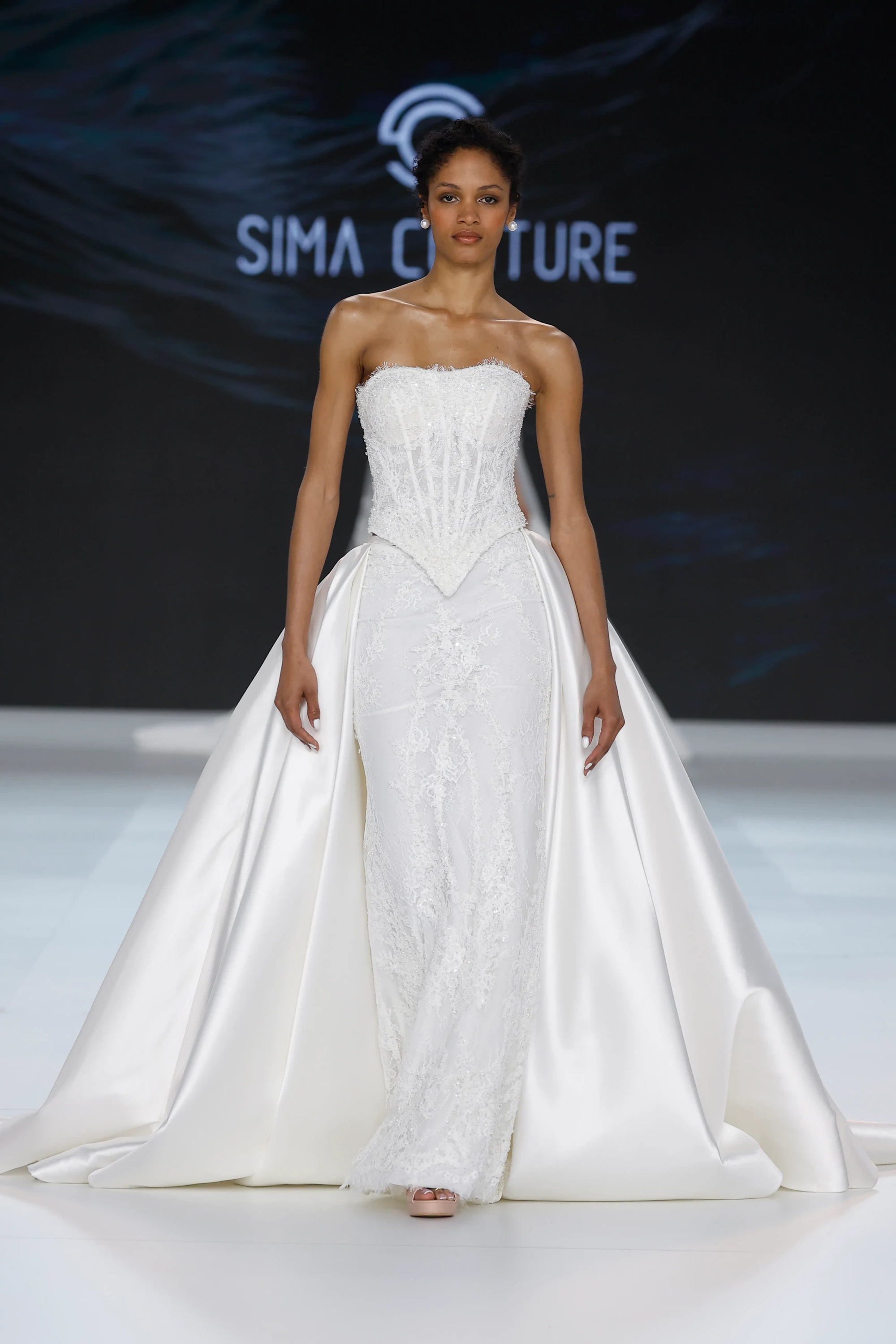 Sima Couture'un Barcelona Bridal Fashion Week 2026 koleksiyonundan bir model, straplez, korsaj detaylı ve ışıltılı dantel işlemelerle süslü, vücudu saran bir gelinlik ve arkadan dramatik biçimde sarkan, parlak saten kumaştan kabarık bir overskirt giyerek 2027 gelinlik trendlerini podyumda sergiliyor.